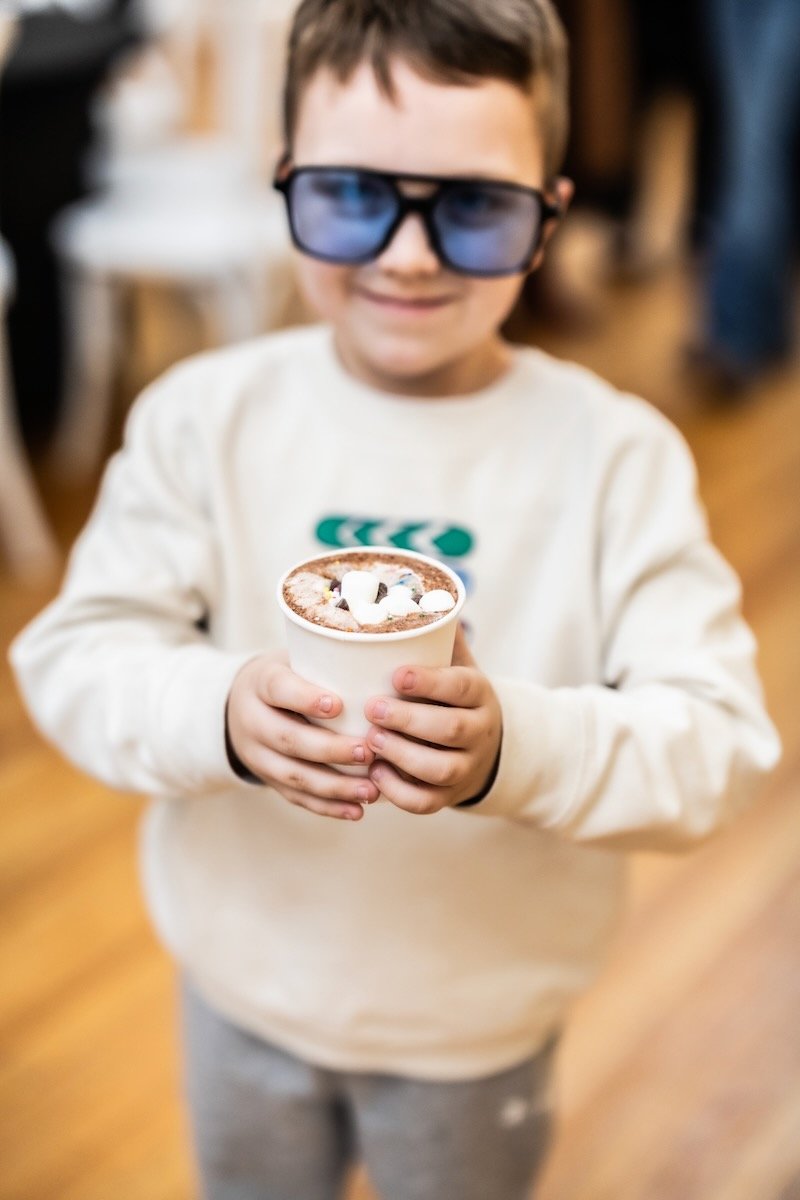 A young boy wearing 3D glasses holding a cup of hot chocolate with marshmallows, smiling indoors.