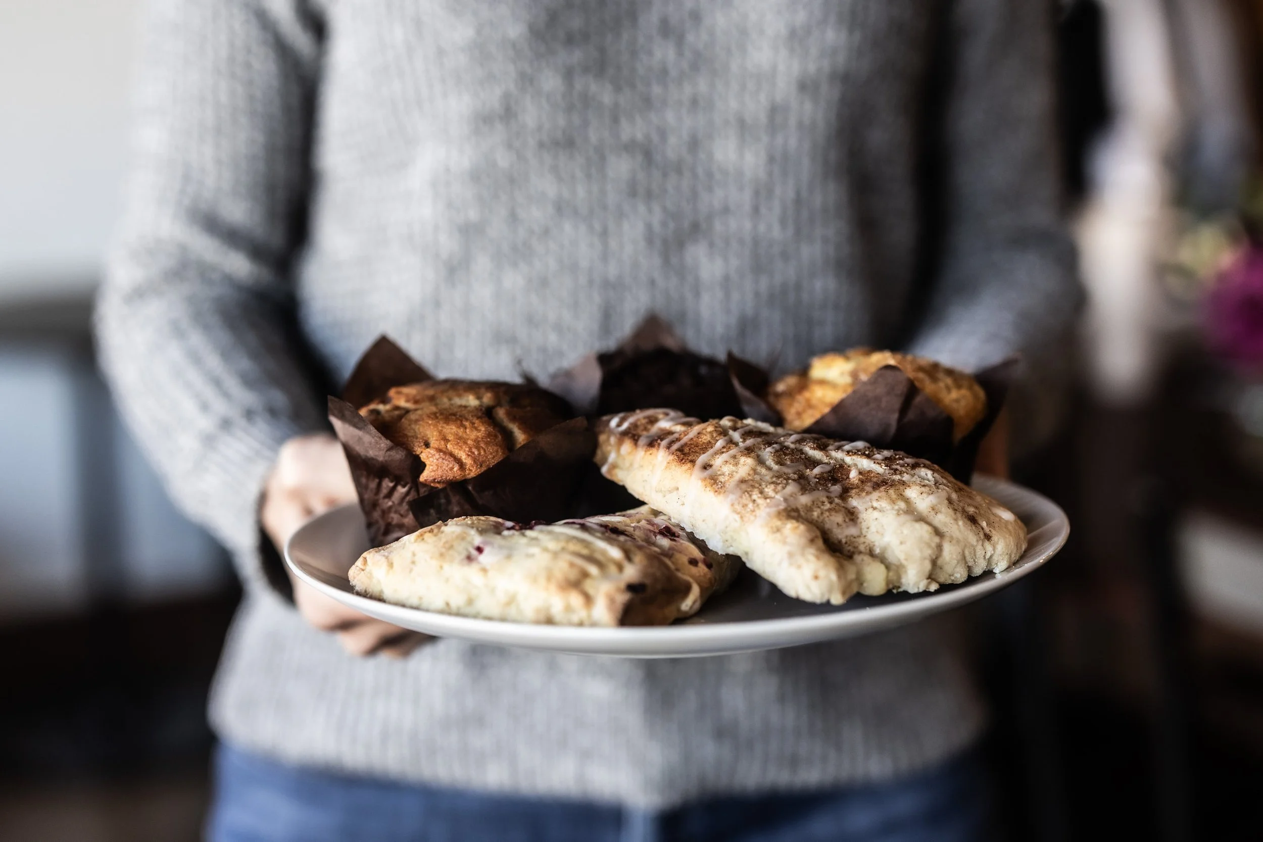 Person holding a white plate with assorted baked goods, including muffins and pastries, in a cozy indoor setting.