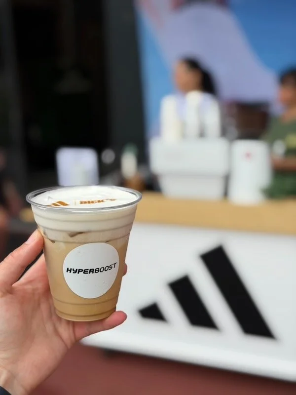 Hand holding a The Coffee Cart iced coffee with 'HYPERBOOST' sticker in front of a blurred background with people working at a The Coffee Cart event in Knoxville and an Adidas logo.