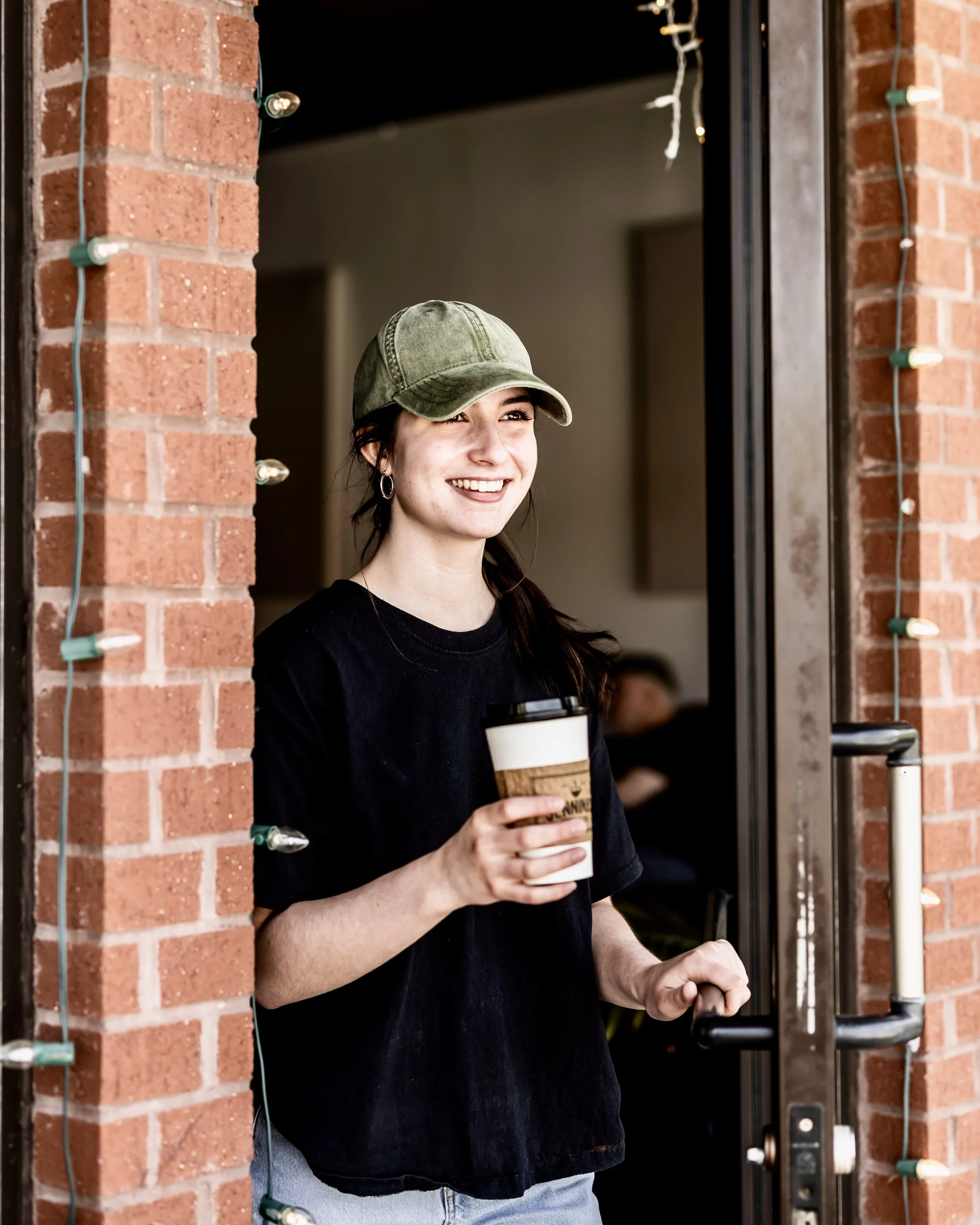A young woman with long dark hair wearing a green cap and black t-shirt, smiling and holding a coffee cup, standing in a doorway decorated with Christmas lights.
