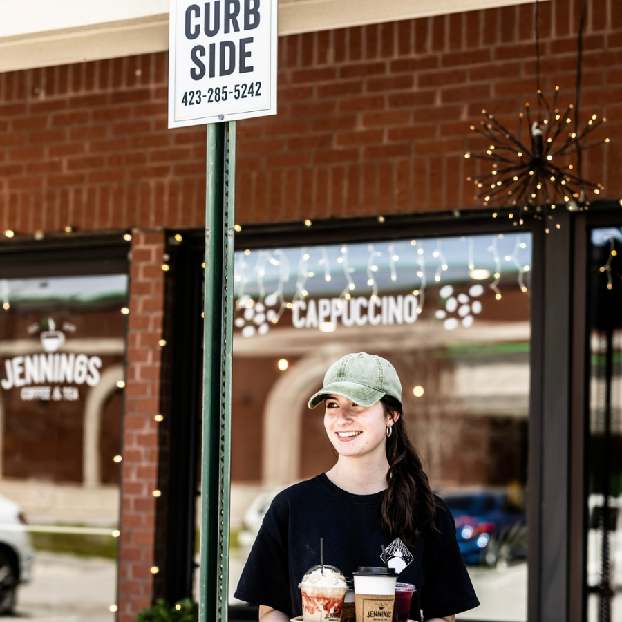 A smiling woman carrying multiple coffee drinks stands outside a coffee shop with string lights, brick walls, and signs reading "Jenninges" and "Cappuccino."