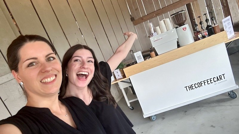 Two smiling baristas taking a selfie in front thier mobile coffee cart branded 'THE COFFEE CART' at a wedding in a barn venue in Knoxville.