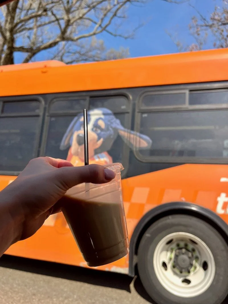A hand holding a plastic cup of iced coffee in front of an knoxville vols orange bus with a cartoon dog mascot on the side.