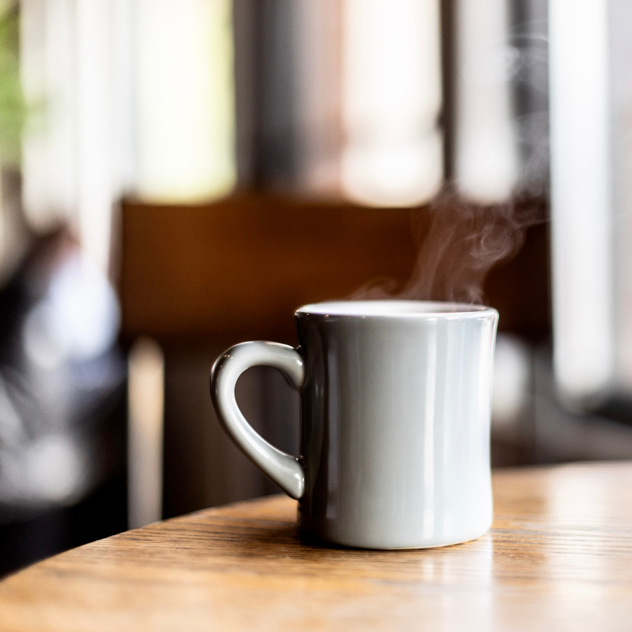 A white ceramic mug filled with a hot beverage, steam rising from it, on a wooden table near a window.