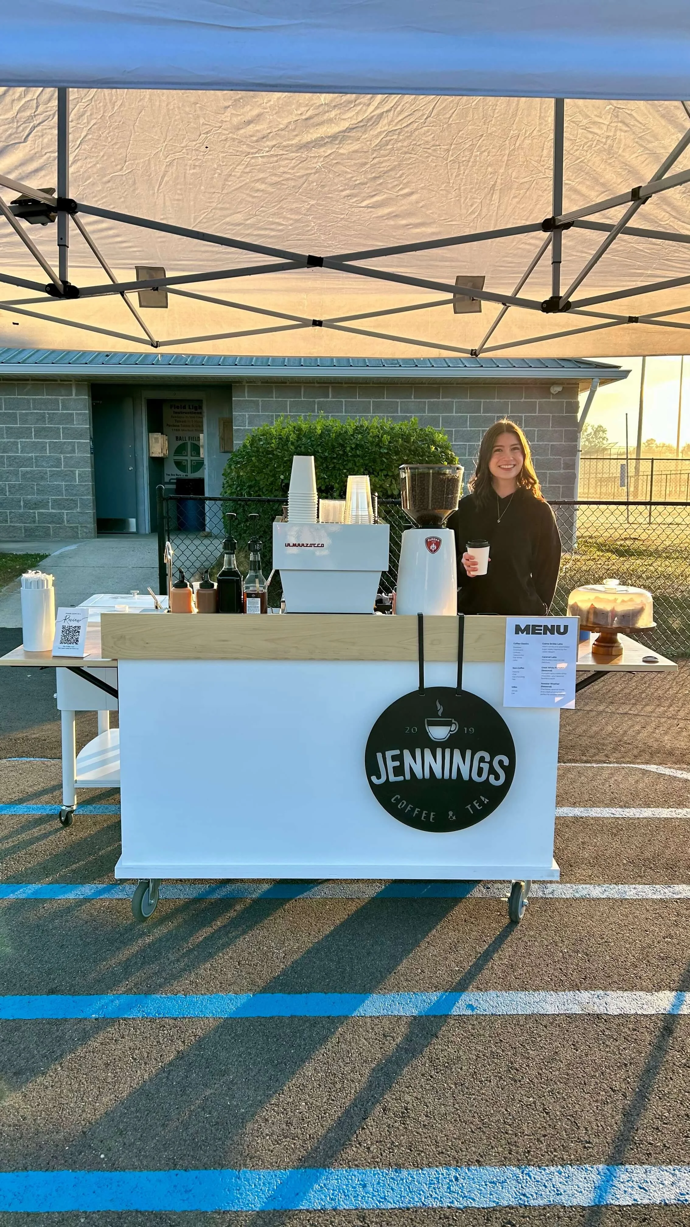 A woman standing behind a coffee and tea stand called Jennings, smiling and holding a coffee cup. The stand is set up outdoors in a parking lot with a canopy overhead, and a building and sunset in the background.