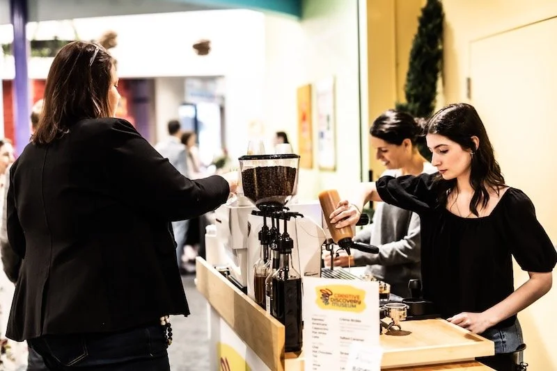 Barista preparing coffee at a counter while a customer waits. Other staff are working behind the counter in a lively café or food court.