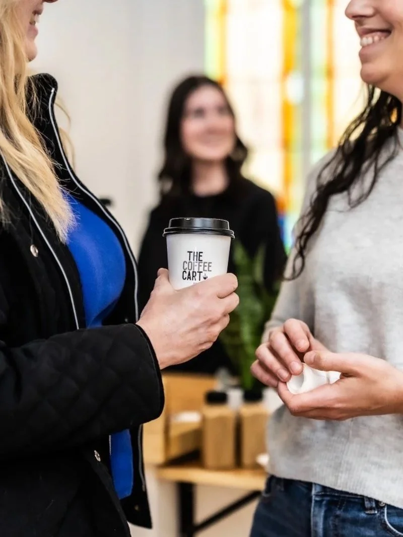 Two women smiling and talking indoors, one holding a white to-go coffee cup with a black lid that reads 'The Coffee Cart'. A person in the background with dark hair and glasses is blurred.