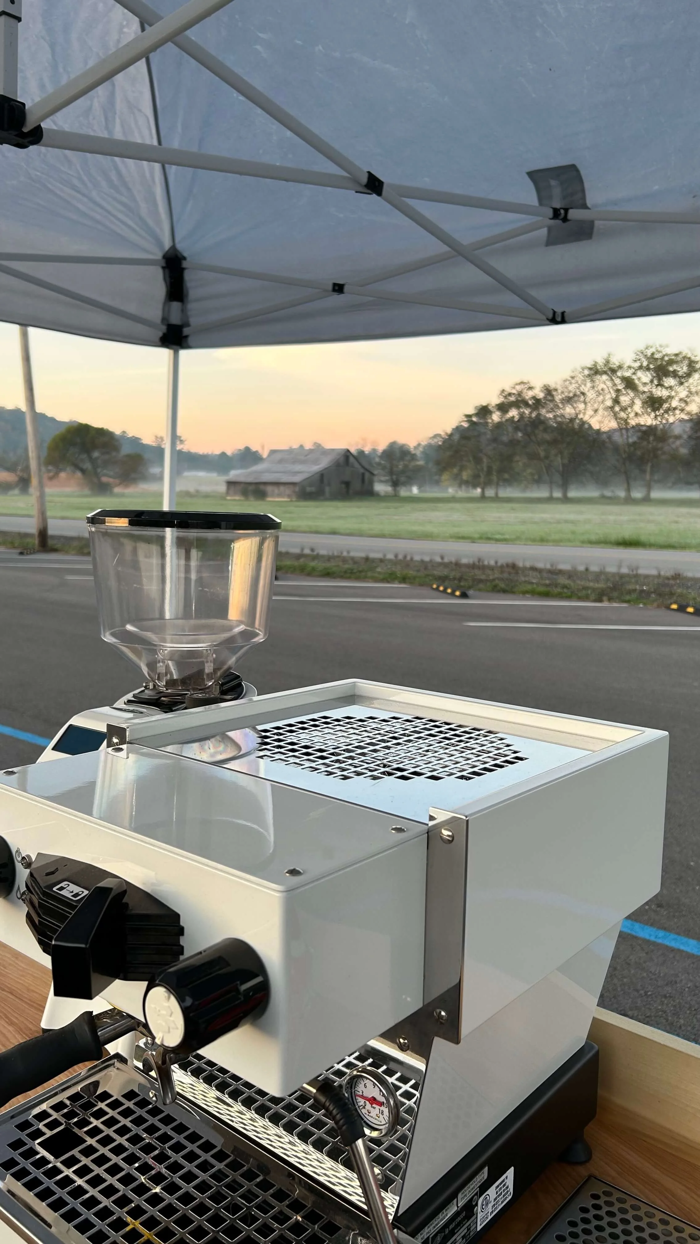 A white espresso machine under a white canopy tent on an outdoor patio, with a view of a rural landscape at sunrise, including trees, a barn, and a road.