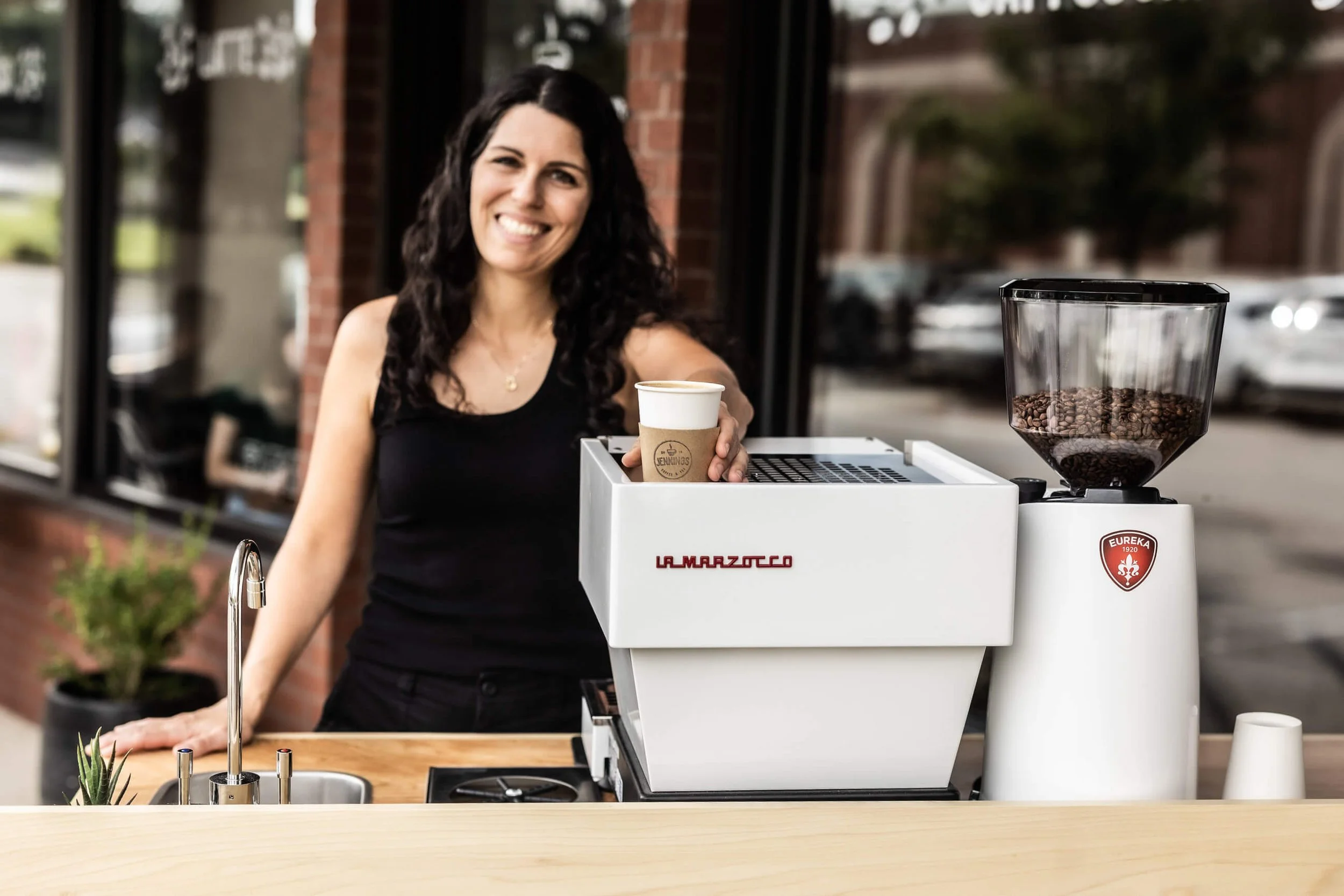 A woman with dark, curly hair handing a coffee cup to a customer at a coffee stand outdoors.