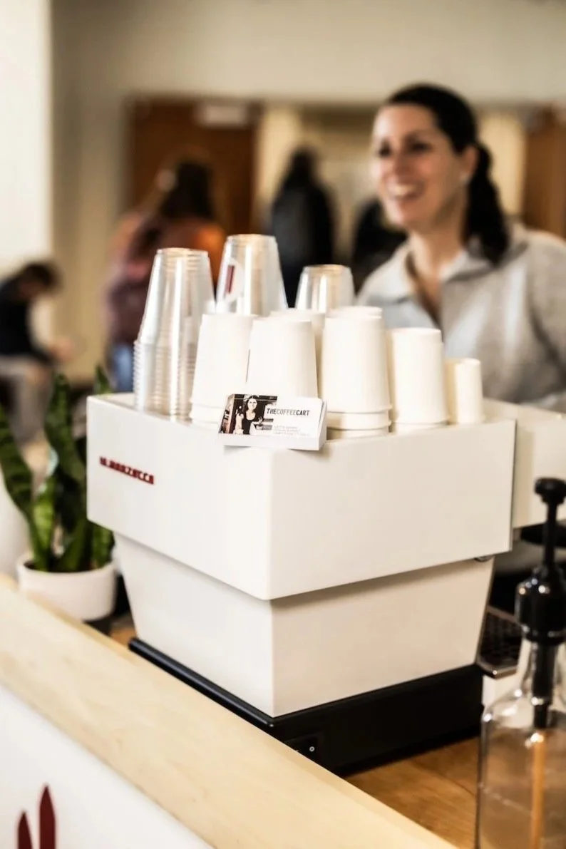 A coffee shop scene showing a white coffee machine with cups and glasses on top, potted plant, and a smiling woman barista in the background.