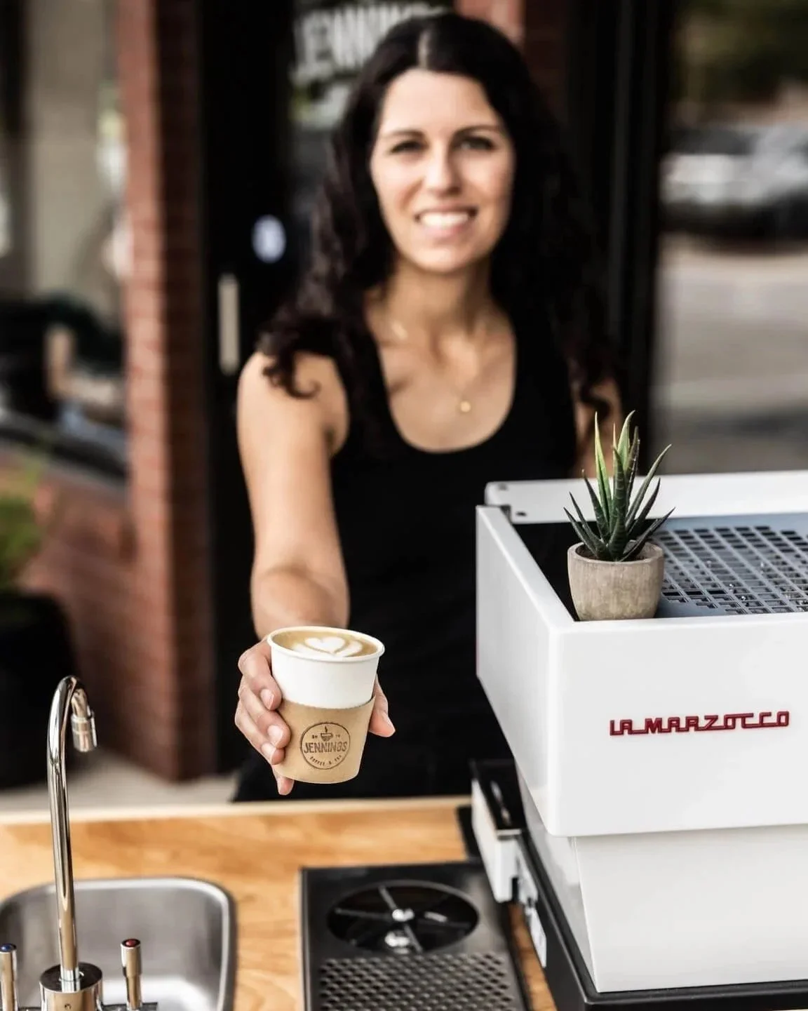 A woman smiling and holding a cup of coffee outdoors at a coffee cart with a potted plant on a white espresso machine.
