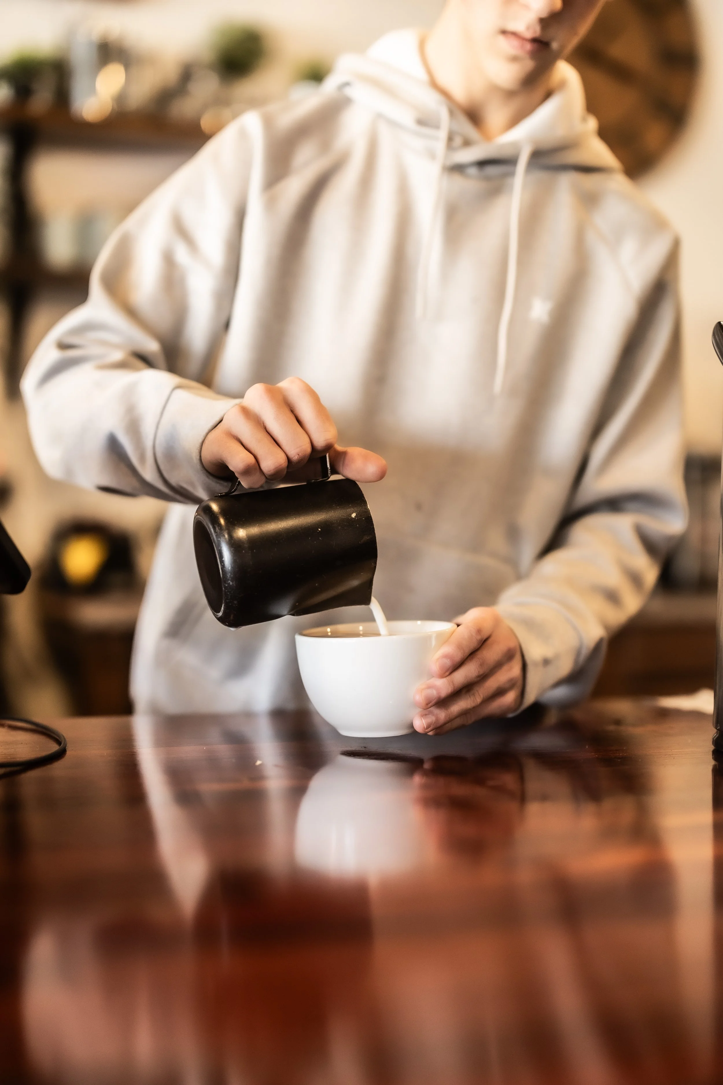 Person pouring milk into a white cup at a wooden counter.
