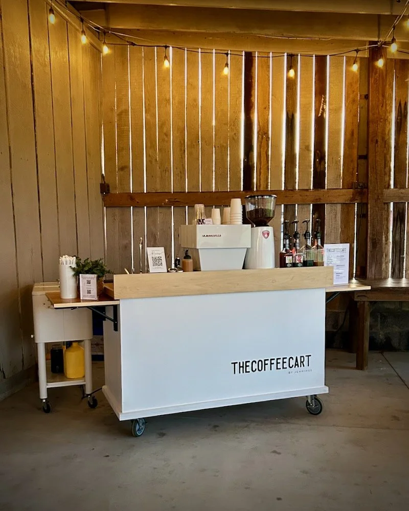 Mobile coffee cart labeled 'TheCoffeeCart' with coffee brewing equipment, cups, and syrup bottles, set against wooden panel walls with string lights overhead. In a wedding venue in Knoxville.
