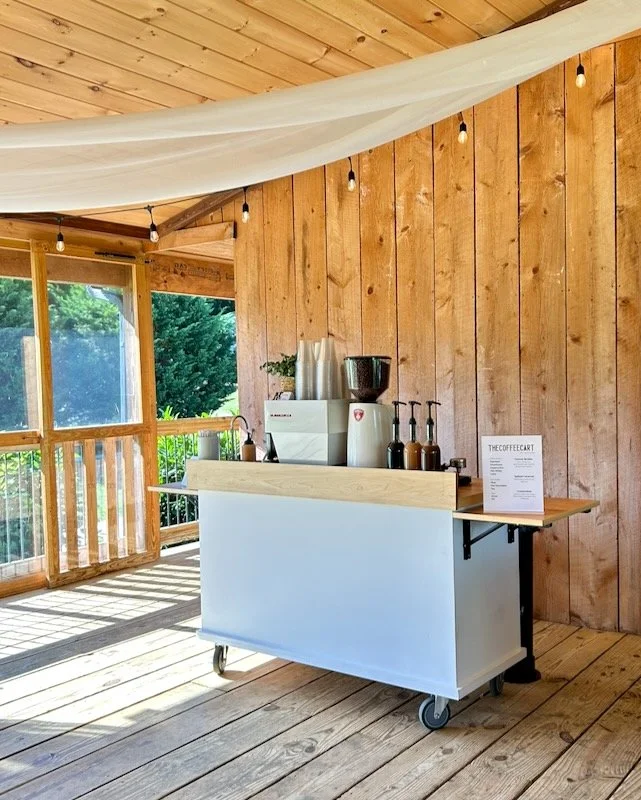 The Coffee cart with espresso machine, coffee beans, and syrups inside a wooden patio at a wedding venue with string lights and a view of green trees outside.