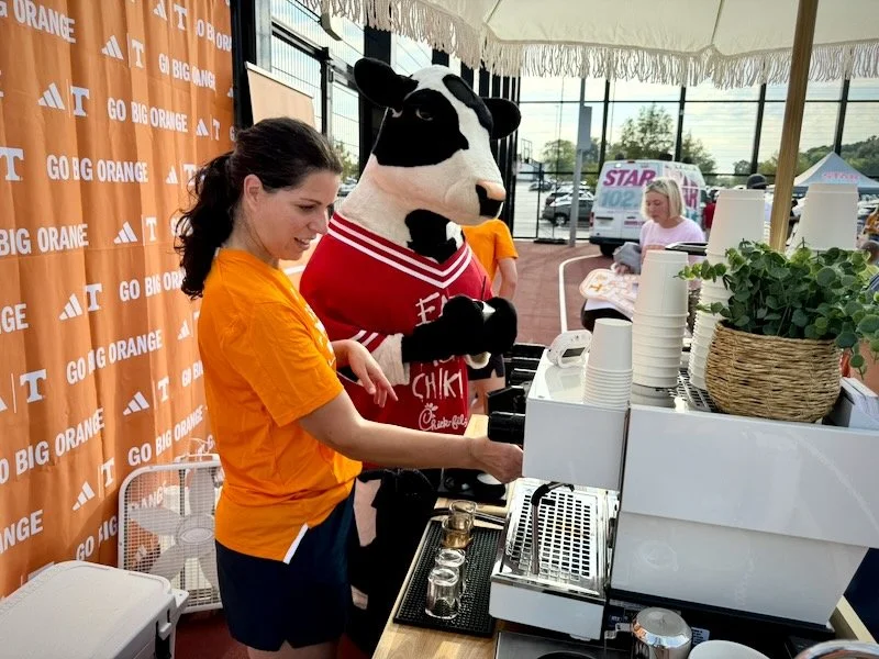 A woman in aorange shirt and a costume mascot of a chick fil-A cow, standing at a The coffee Cart. The background features a promotional Tennessee orange backdrop with the University of Tennessee logo and several people in the background.