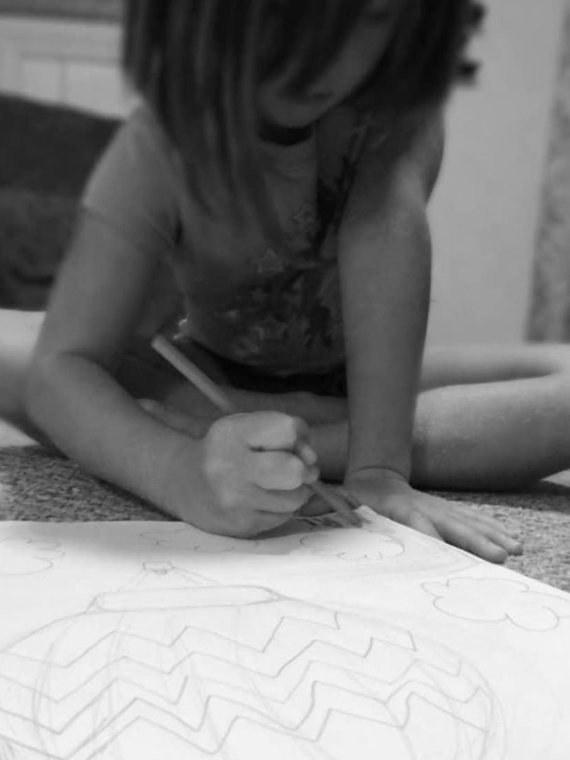 Child sitting on the floor drawing or writing on a large sheet of paper with zigzag patterns, using a crayon or pen, in a room with carpeted floor.