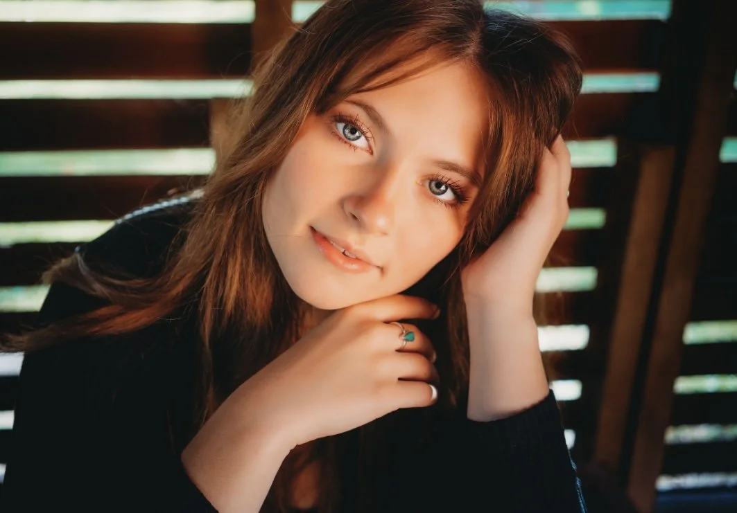 A young woman with long reddish-brown hair and blue eyes, resting her chin on her hand and tilting her head to the side, indoors with wooden slats in the background.
