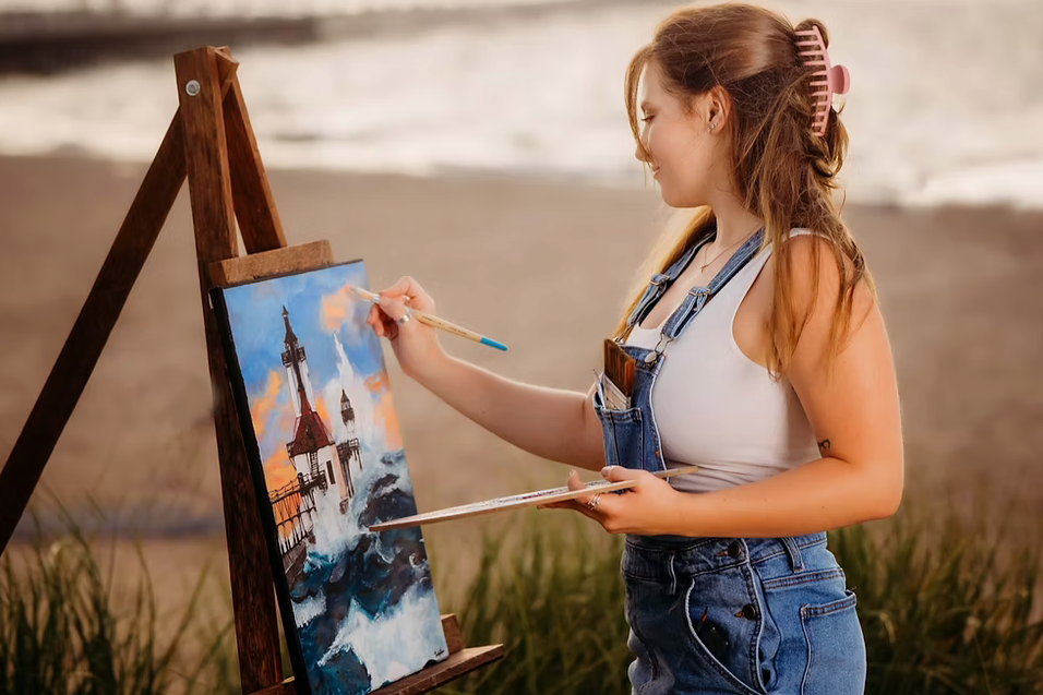 Young woman painting a seascape on an easel at the beach, wearing denim overalls and a white tank top, with her hair in a pink hair clip.