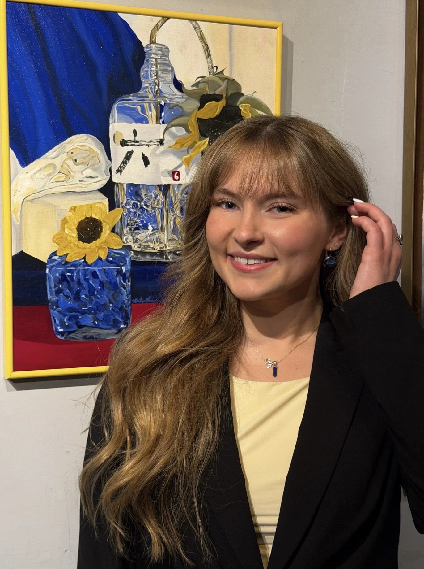 Young woman with long wavy hair, smiling, wearing a black blazer and a light-colored top, standing in front of a colorful still life painting of a sunflower in a blue vase, a bar of soap, a bottle, and a sunflower, displayed on a wall.