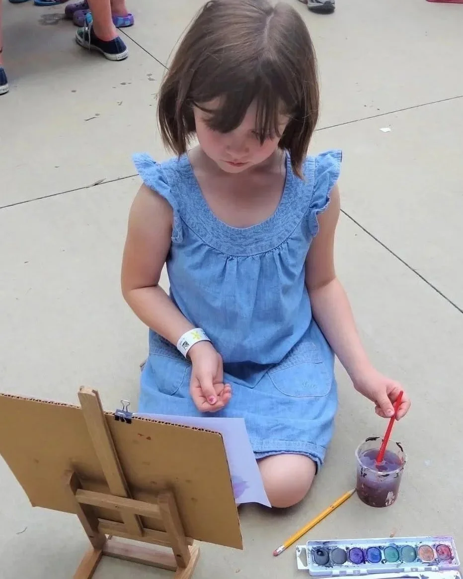 A young girl kneeling on the concrete floor, painting on paper set on a small wooden easel. She is wearing a blue dress and has a watercolor paint set and a cup of water with a red paintbrush in it in front of her.