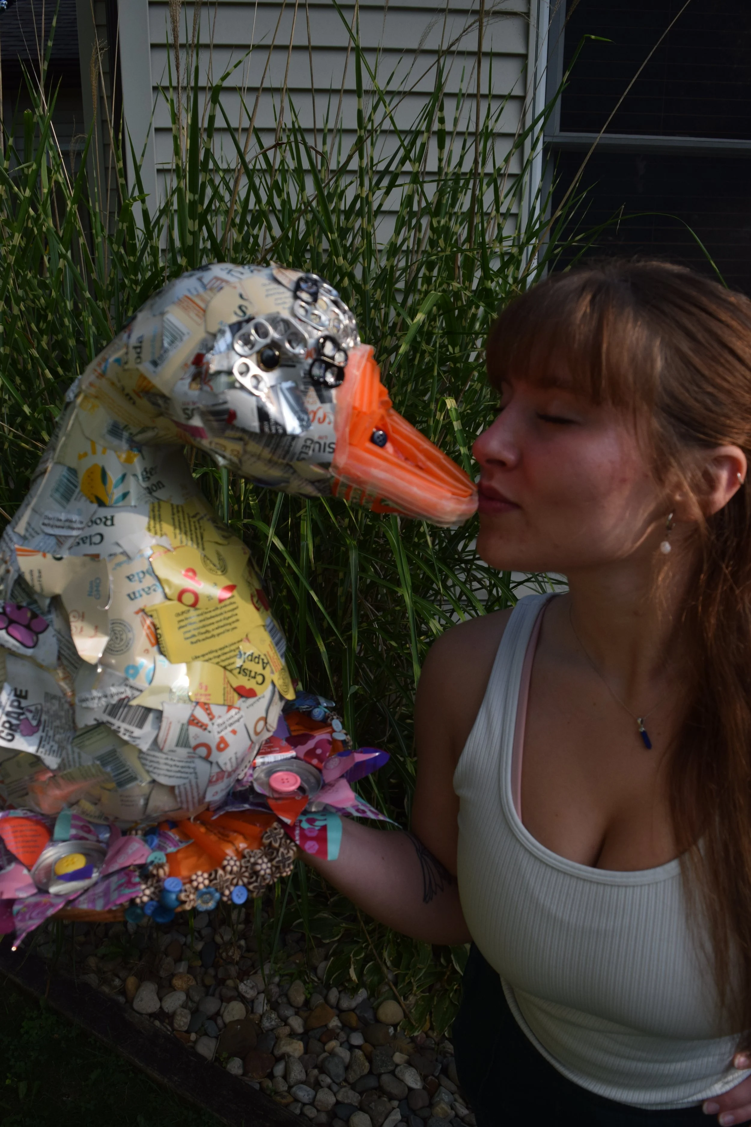 A woman with brown hair wearing a white tank top and earrings, is about to kiss a sculpture of a snake made of crumpled paper and metal, outdoors next to a house with siding and tall grass.