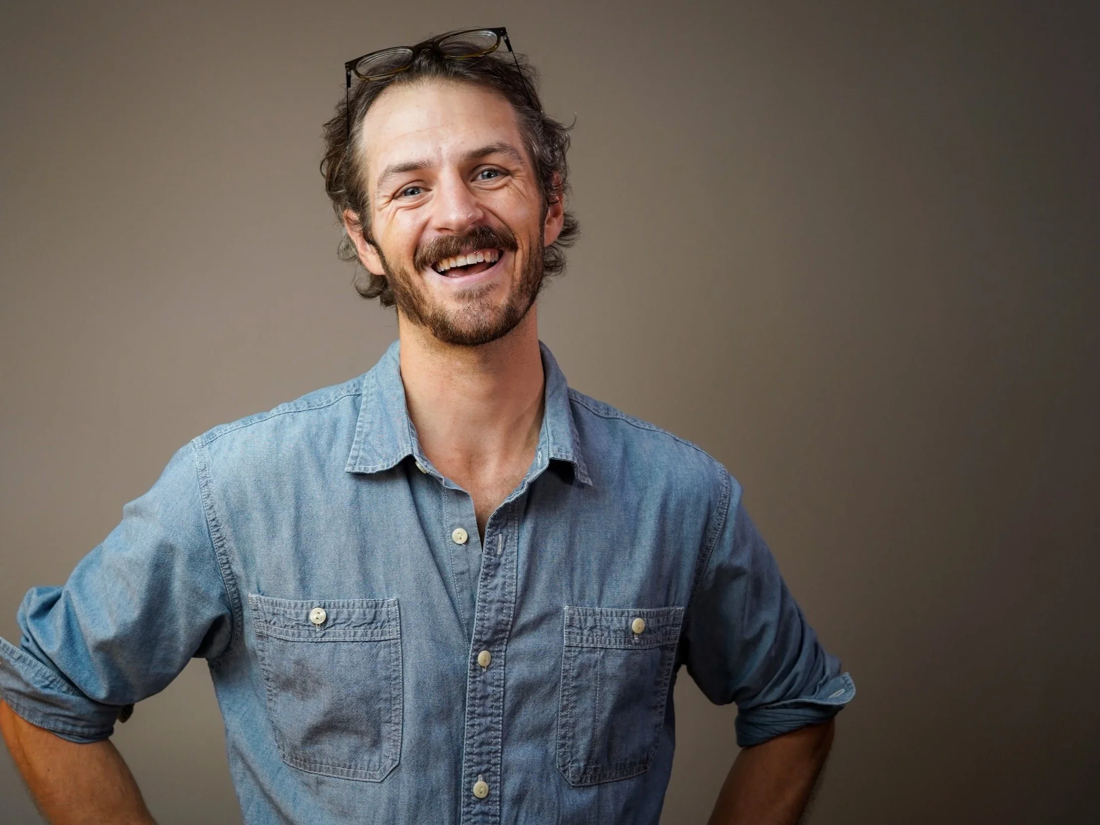 A smiling man with a beard and glasses on his head, wearing a denim shirt, standing against a neutral background.
