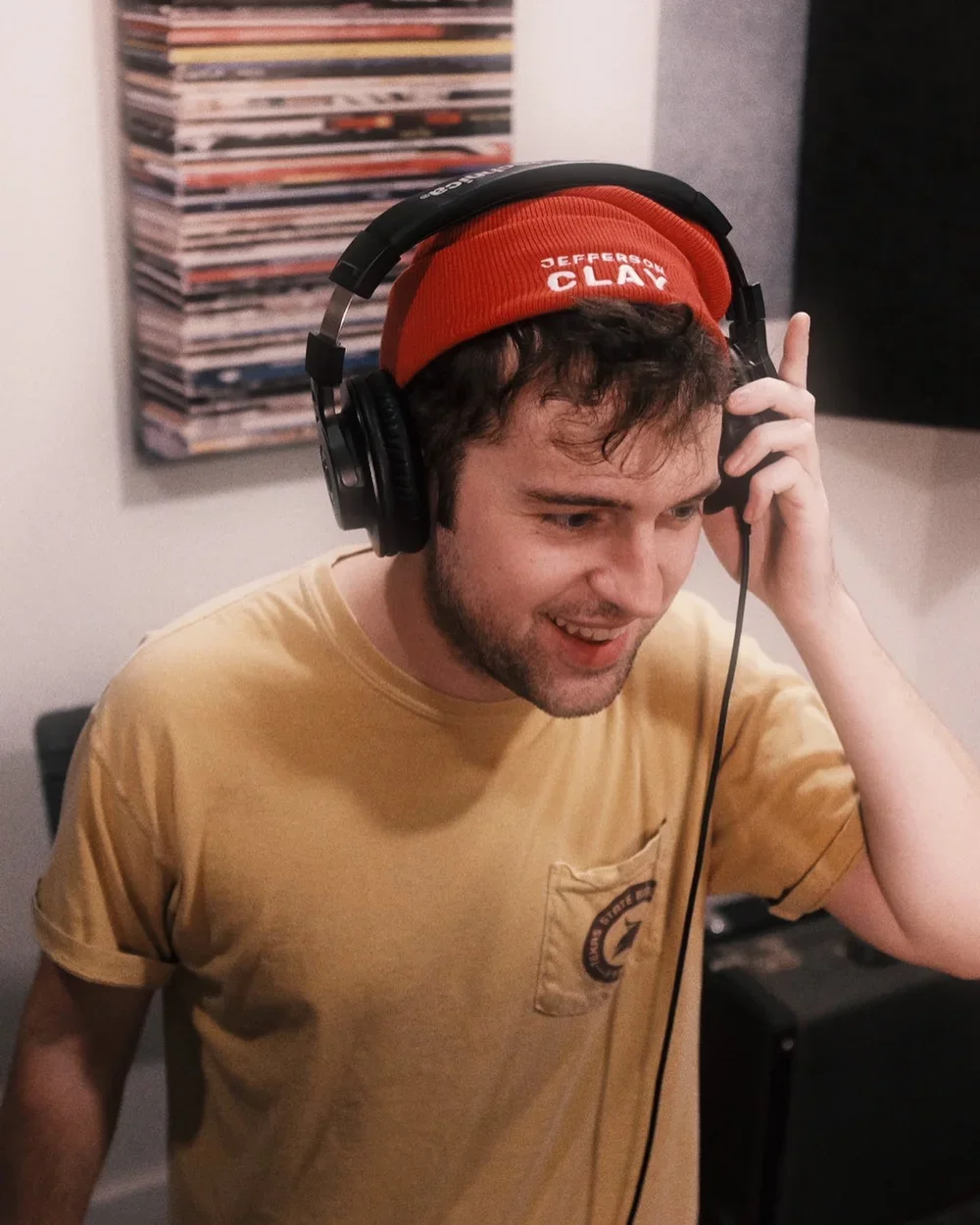 Young man with curly brown hair, wearing a red beanie hat labeled 'Jefferson Clay', headphones, and a beige t-shirt, smiling and listening to music in a room with a wall decorated with acoustic panels and stacked records or magazines in the background.