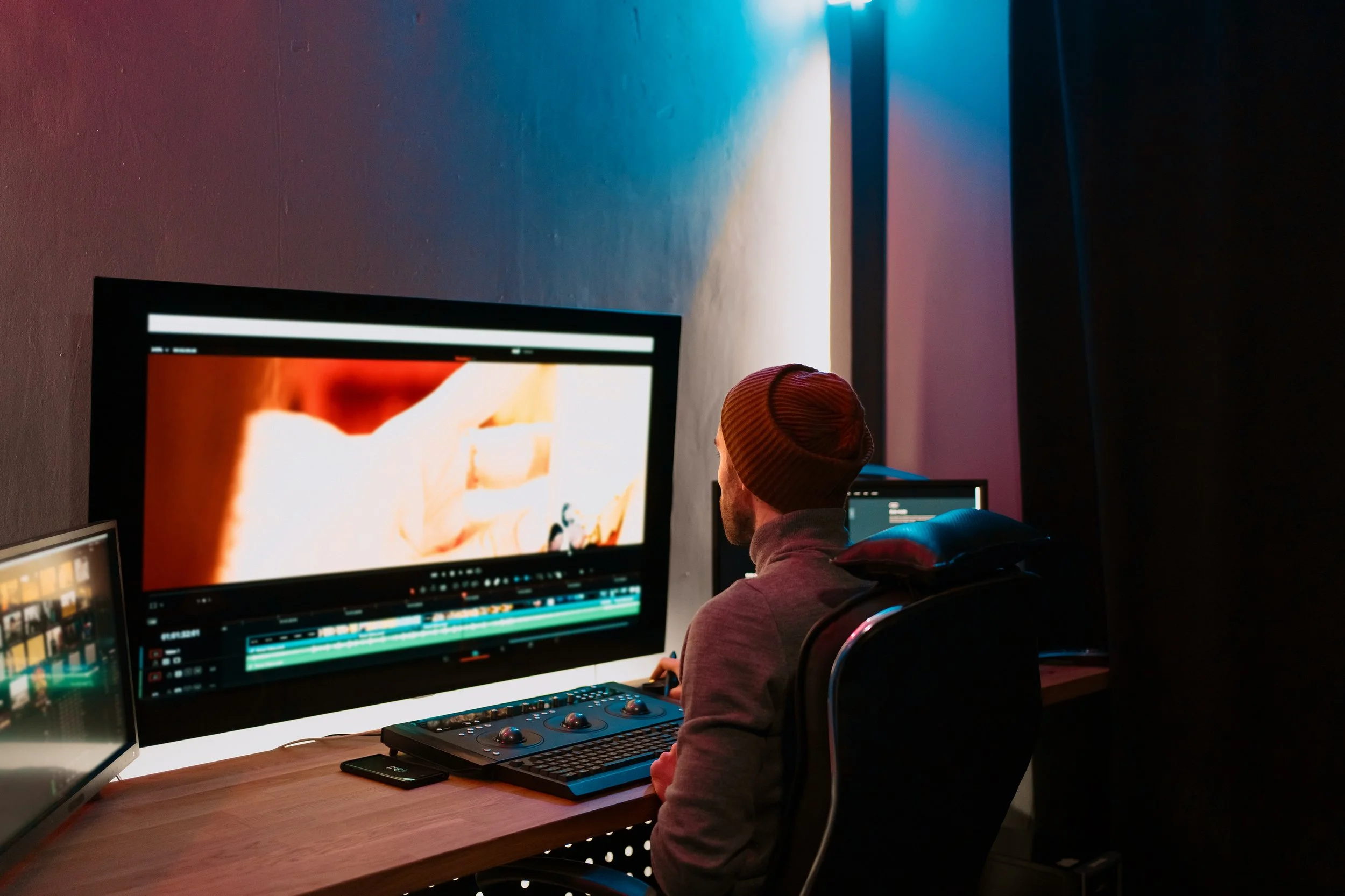 A man wearing a beanie and hoodie sitting at a desk with multiple monitors, editing a video in a dimly lit room.