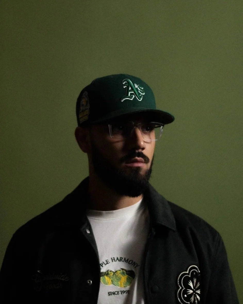 A man wearing glasses, a green cap, and a black jacket standing against a green wall.