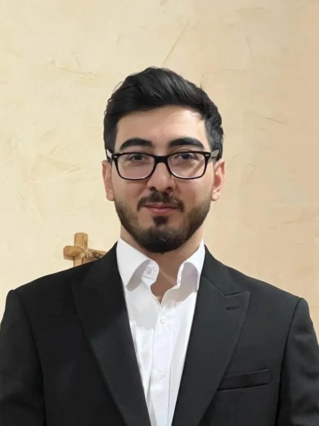 A young man with dark hair, glasses, and a beard wearing a formal black suit and white shirt standing against a beige wall.