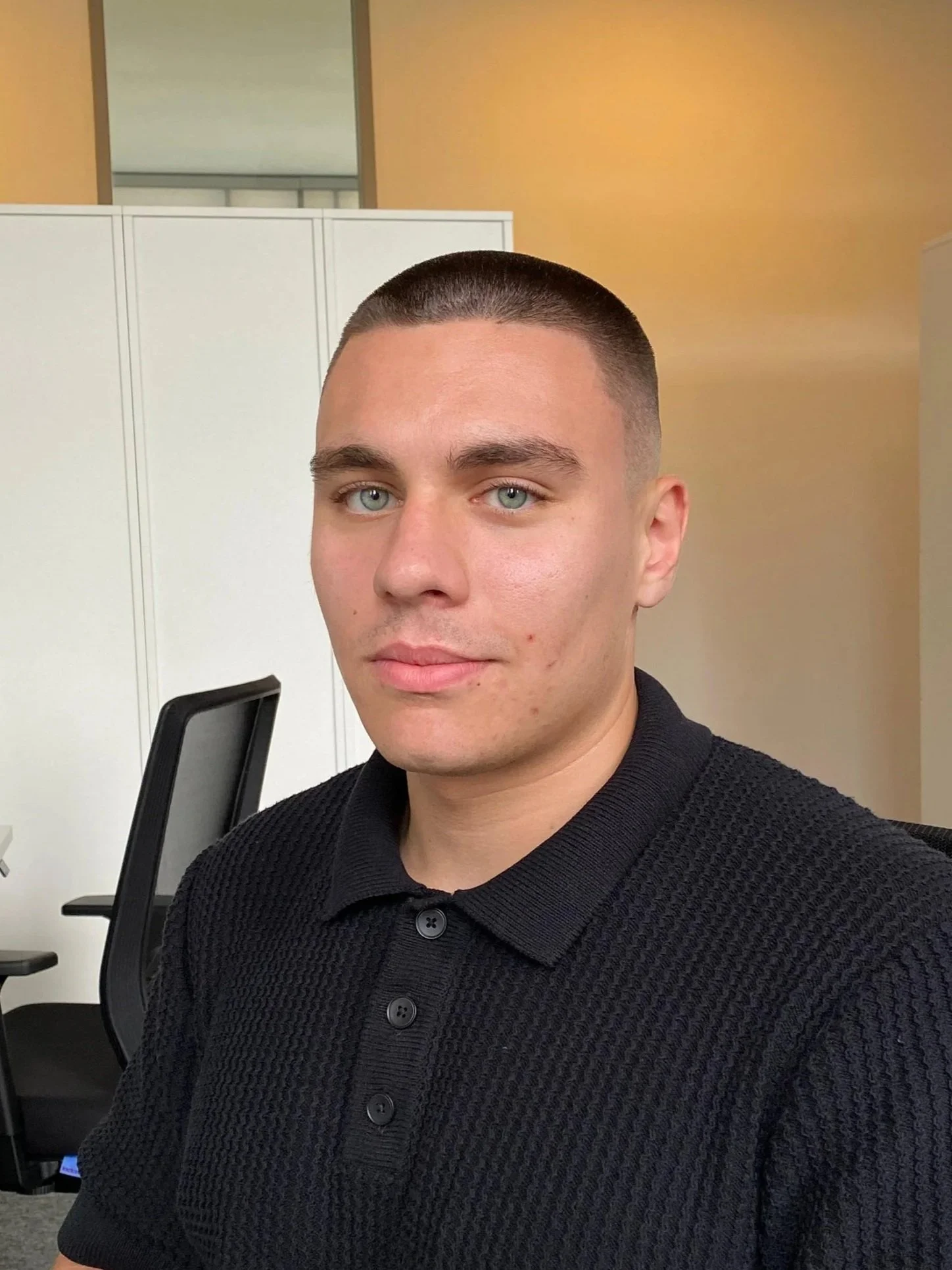A young man with short dark hair and blue eyes takes a selfie in an office setting, wearing a black textured polo shirt, with office chairs and white cabinets in the background.