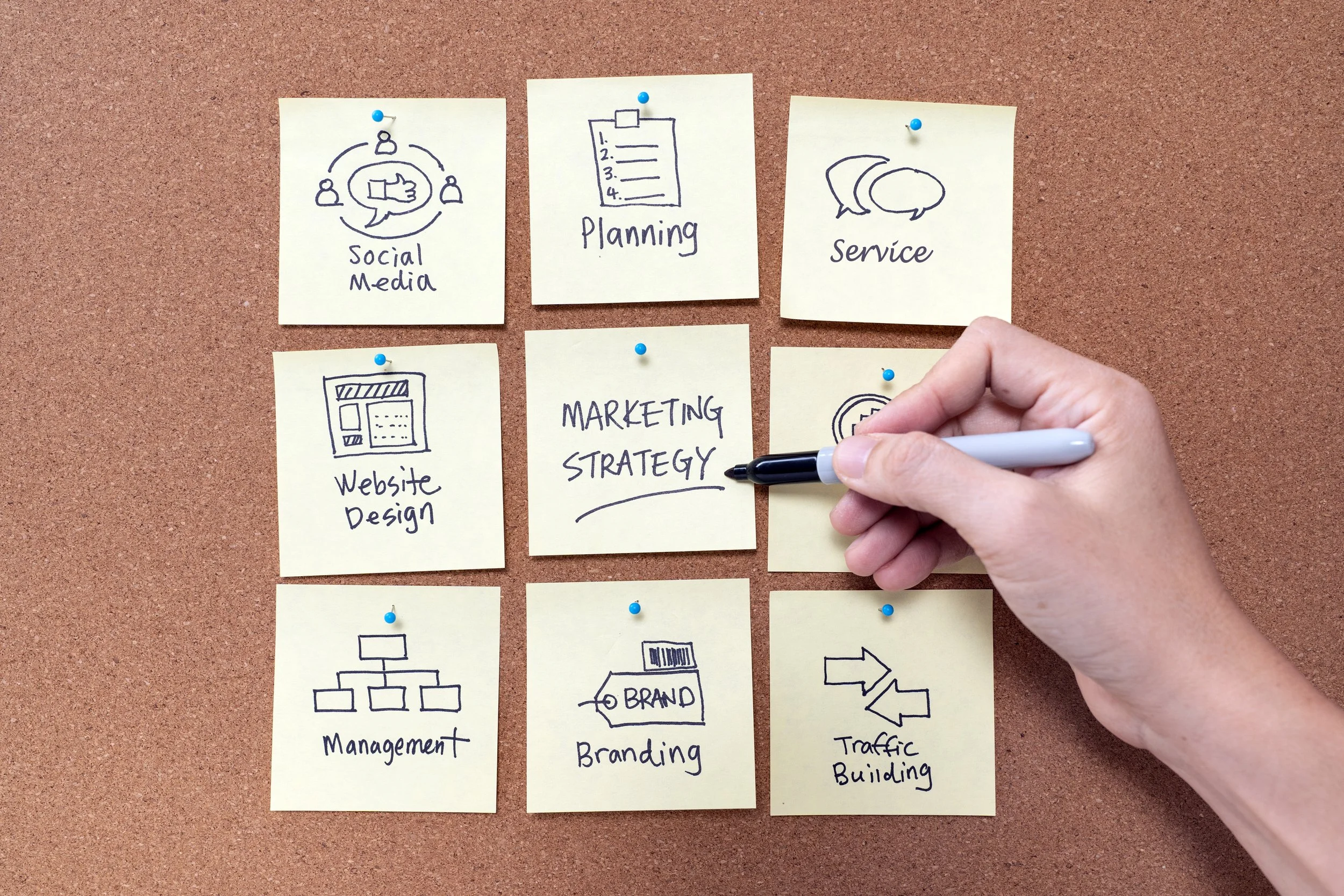 Hand writing 'Marketing Strategy' on a sticky note surrounded by other sticky notes with marketing-related icons and words, arranged on a corkboard.