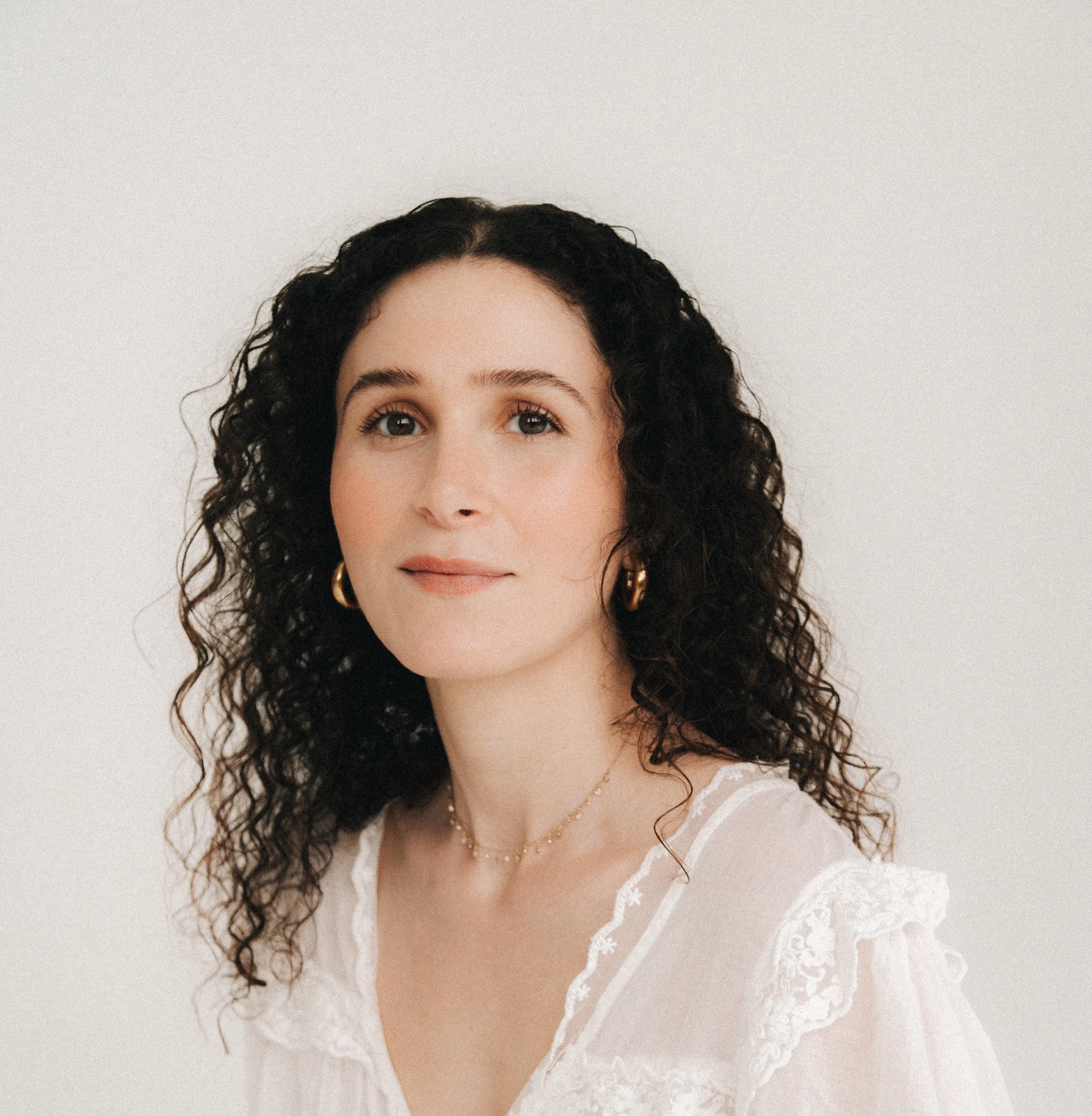 A woman with dark curly hair, wearing gold hoop earrings, a delicate gold necklace, and a white lace blouse, standing against a plain white background.