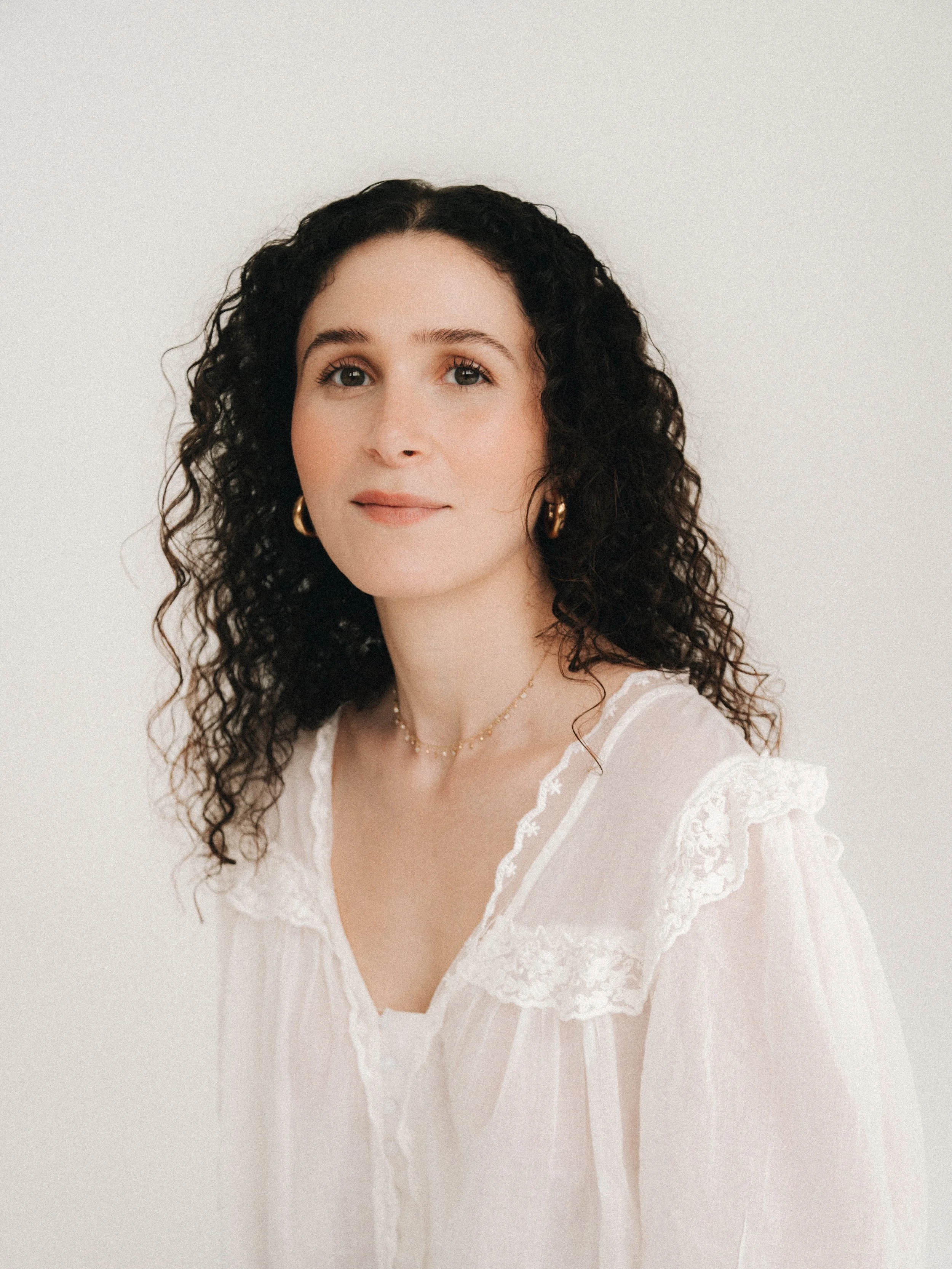 A woman with curly dark hair wearing a cream lace blouse, gold hoop earrings, and a delicate necklace, posing against a plain light background.