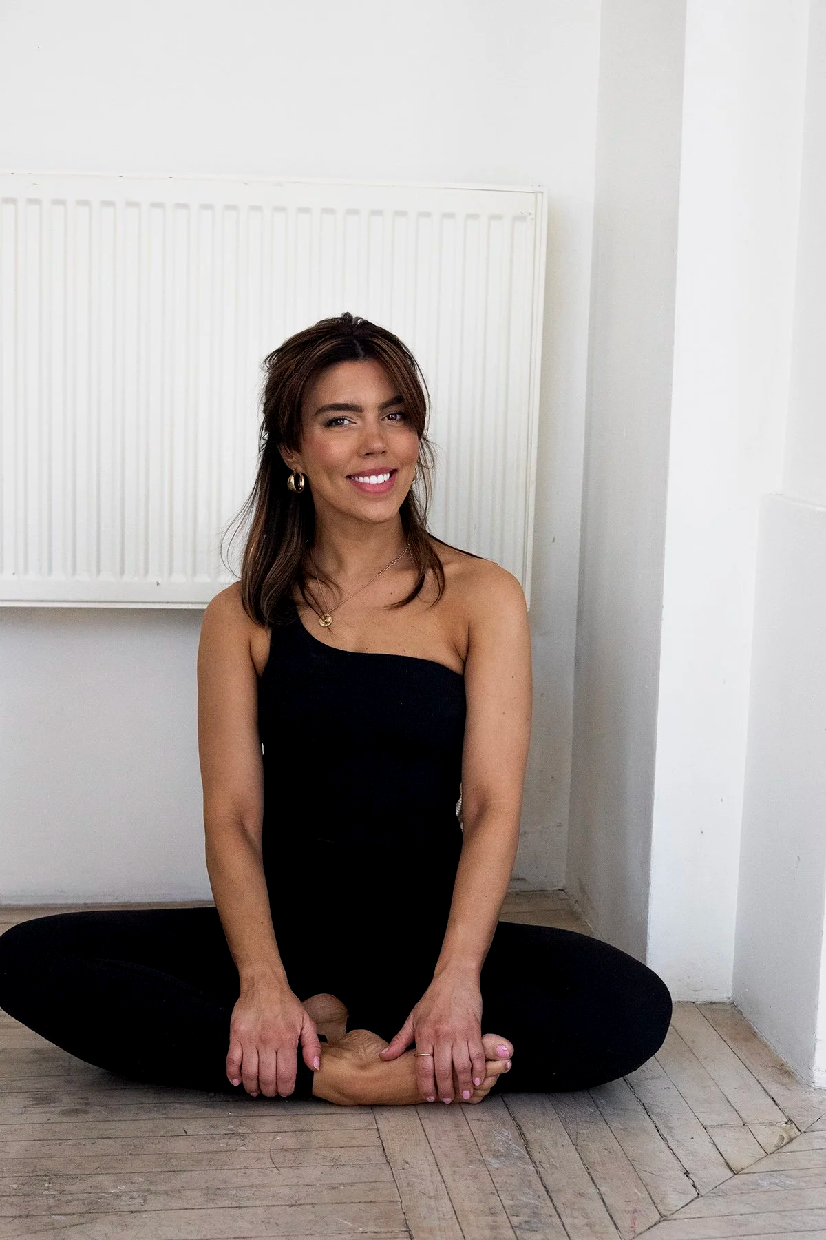 A woman sitting cross-legged on the floor wearing a black strapless top and black pants, smiling at the camera in a minimal white-walled room.