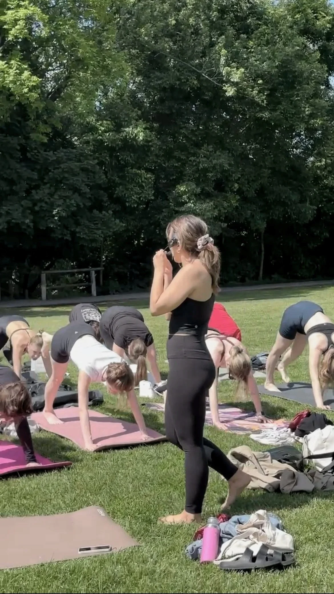 A group of people participating in an outdoor yoga class on a grassy area with trees in the background. There is a woman standing in the foreground, observing or preparing to join the class.