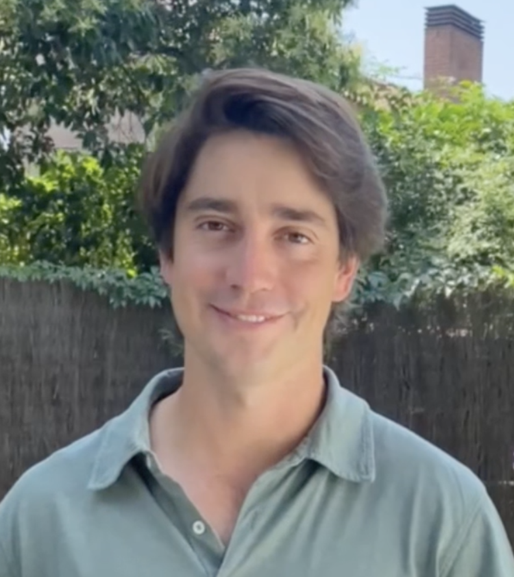 Joven sonriendo con cabello castaño oscuro, lleva una camisa de color gris claro, en un fondo de árboles y un muro de madera.