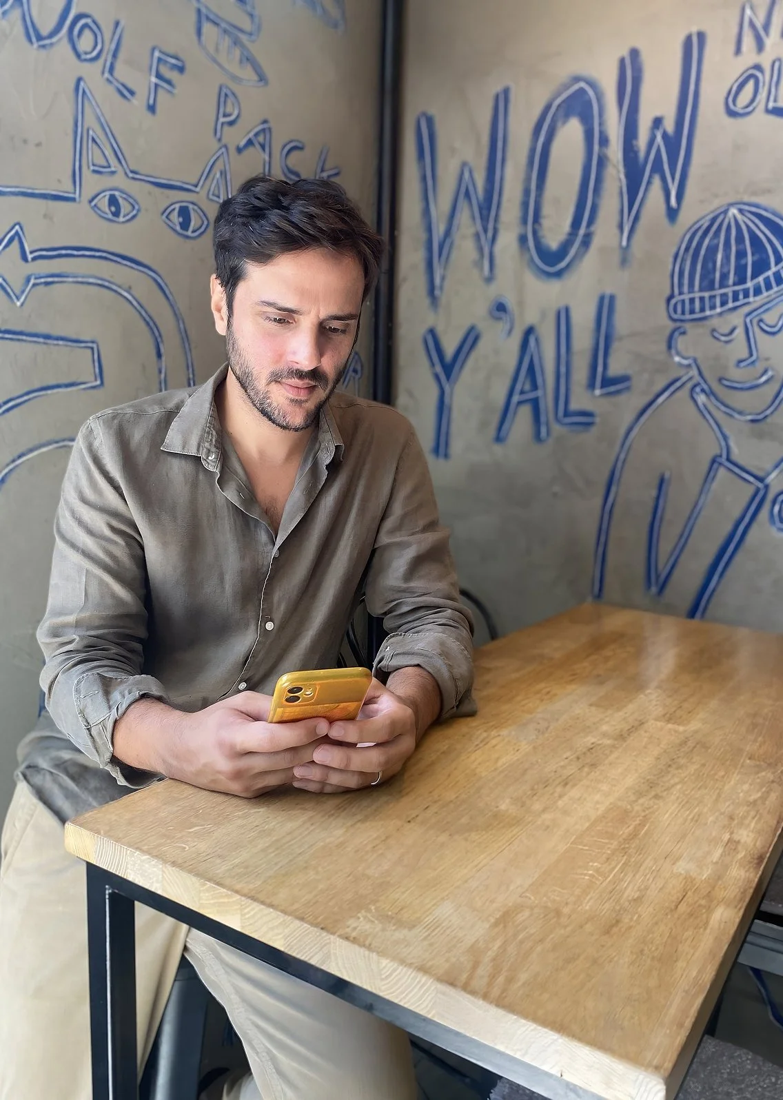 Alex Gallardo, de Brand Mentor, sentado en una mesa de madera en un café, mirando su teléfono móvil color amarillo, con paredes decoradas con dibujos de ojos, textos como 'WOW' y 'Y'ALL' en el fondo.