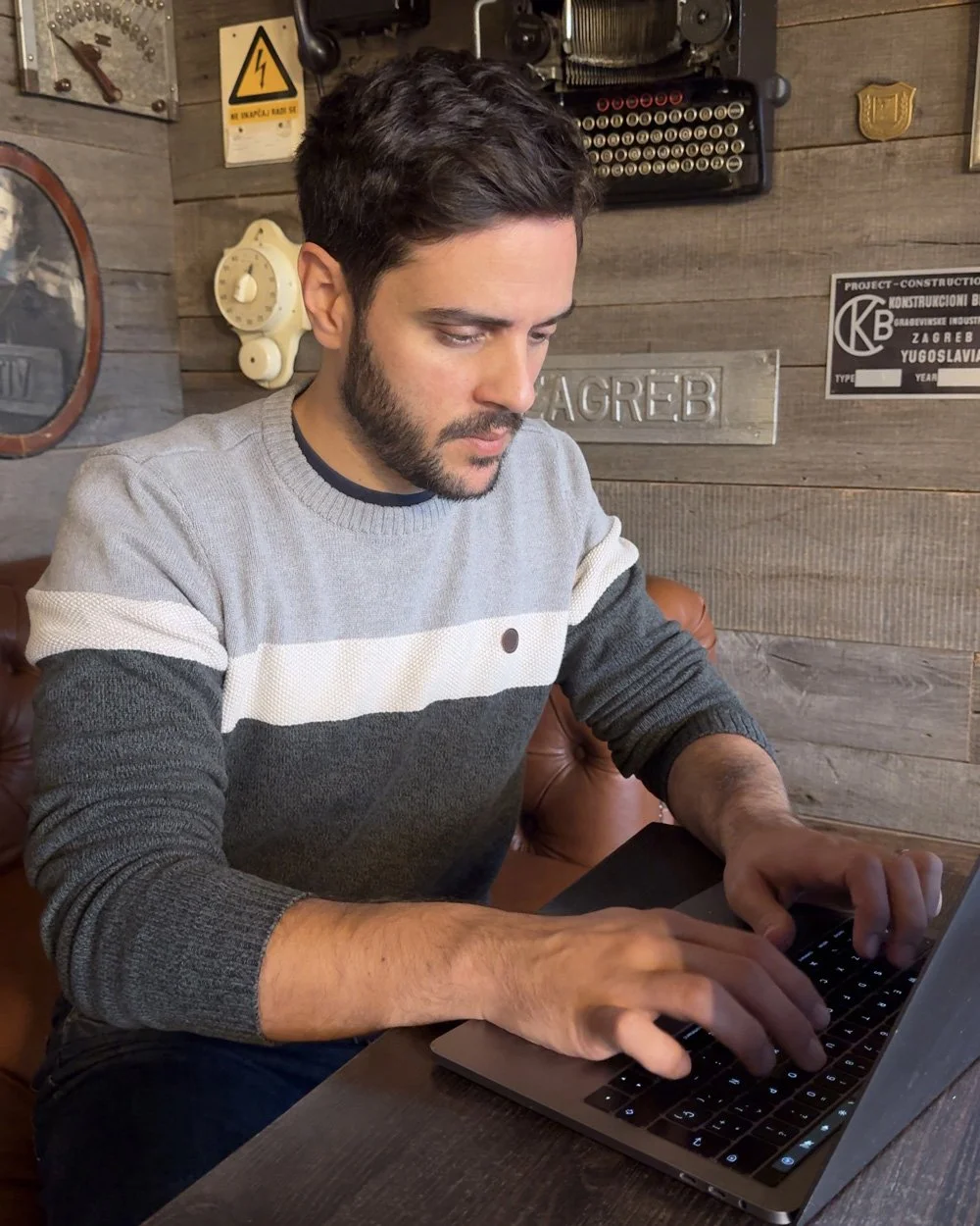 Alex Gallardo, de Brand Mentor, sentado en una mesa de madera, usando una laptop en una habitación con paredes de madera, decorada con signos y objetos vintage.