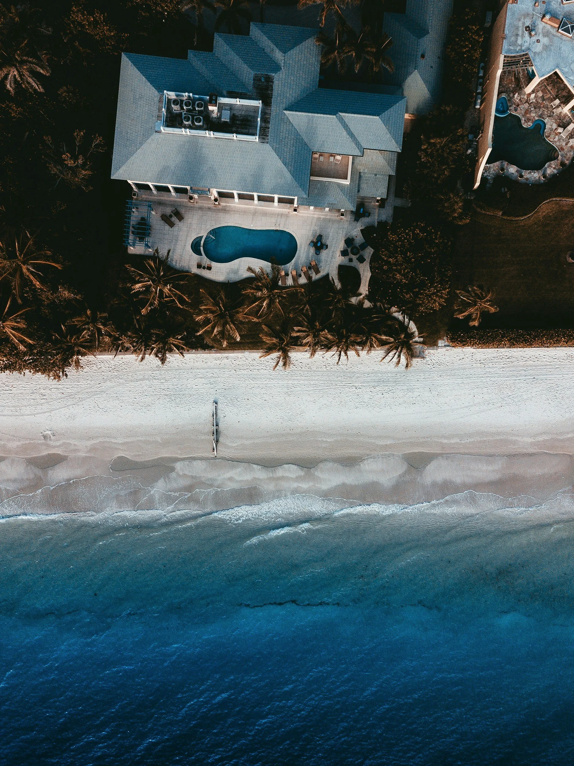 Aerial view of a house with a swimming pool, surrounded by palm trees, adjacent to a beach with ocean waves.