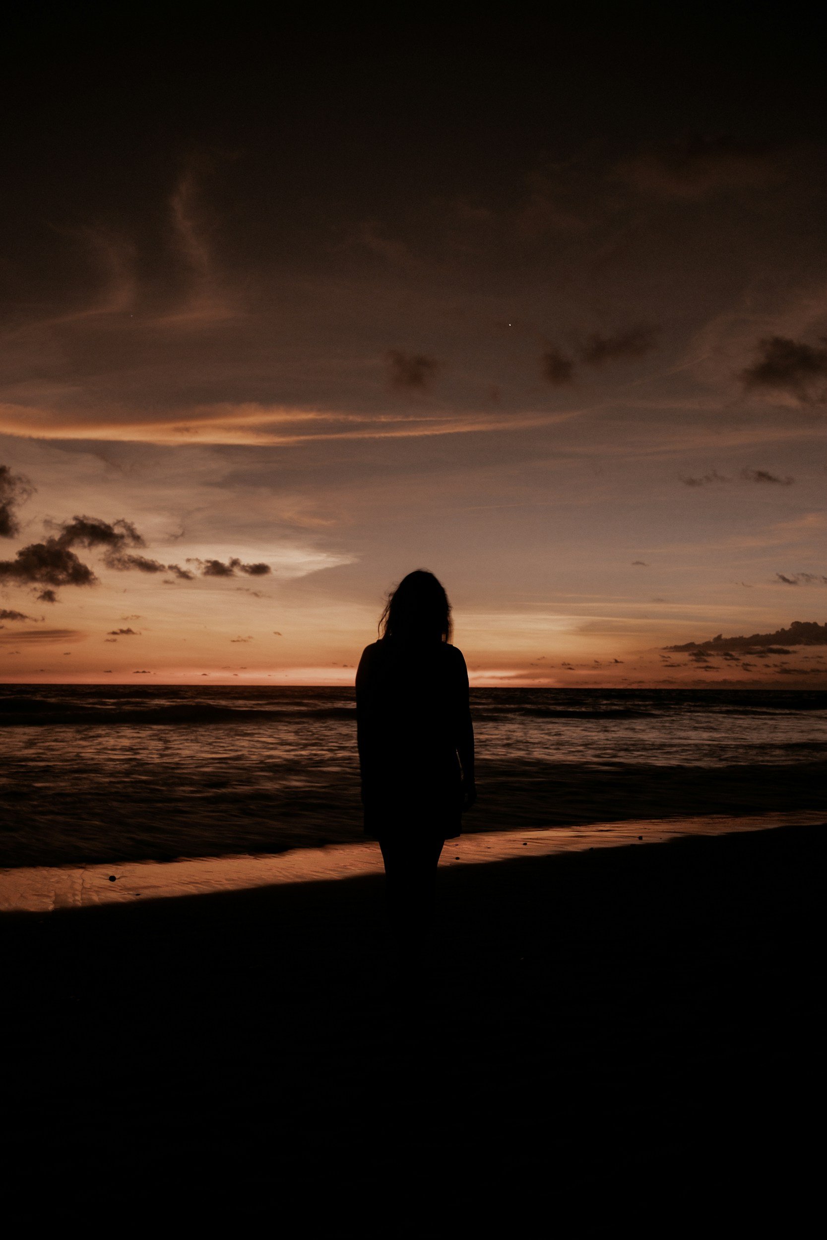 Silhouette of a woman standing on the beach at sunset, with colorful sky and ocean waves in the background.