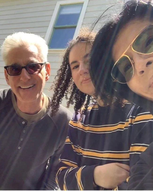 Three people taking a selfie outdoors in front of a light-colored building with a window. The group includes an older man with white hair wearing sunglasses and a gray shirt, a woman with curly hair in a striped shirt, and another woman with dark hair and sunglasses partially visible.
