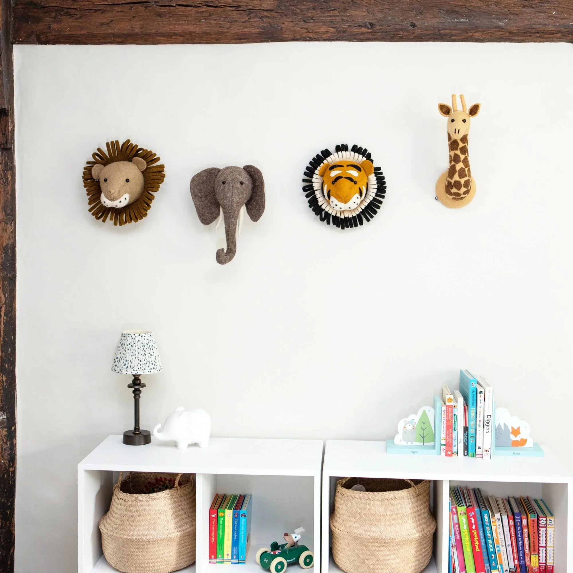 Wall with four animal plush heads: lion, elephant, tiger, and giraffe, above two white storage units with books, baskets, a lamp, and a decorative toy.