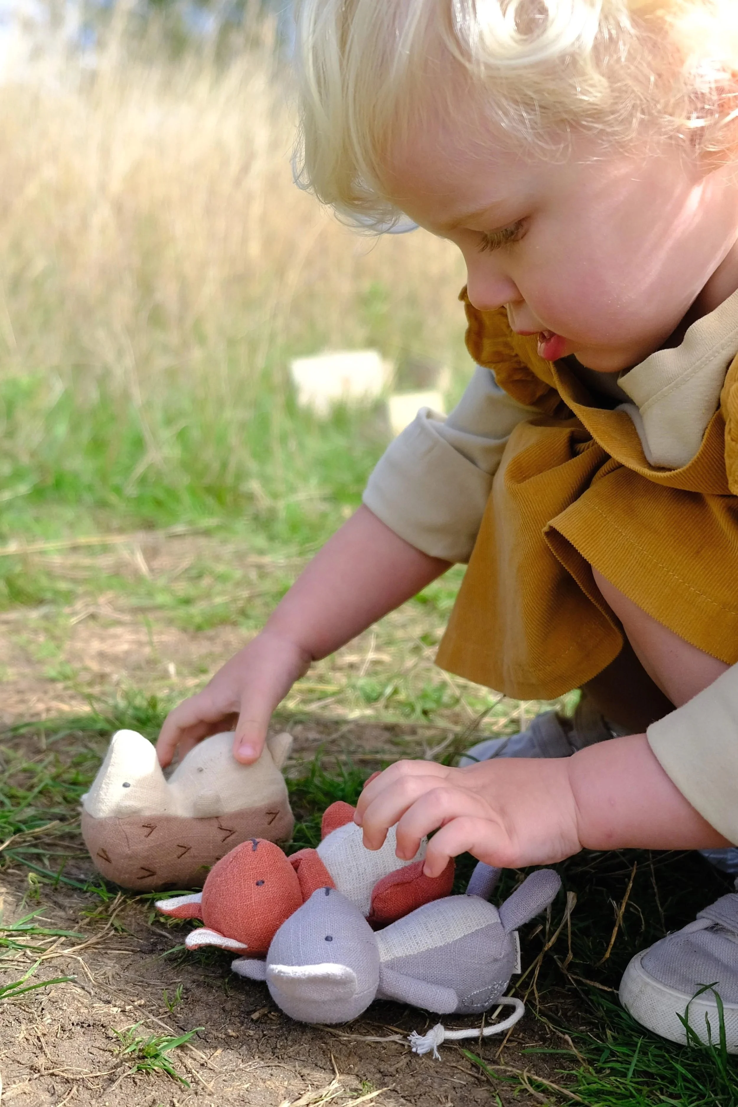 A young child playing outdoors on the ground with stuffed animal mice in a grassy field.