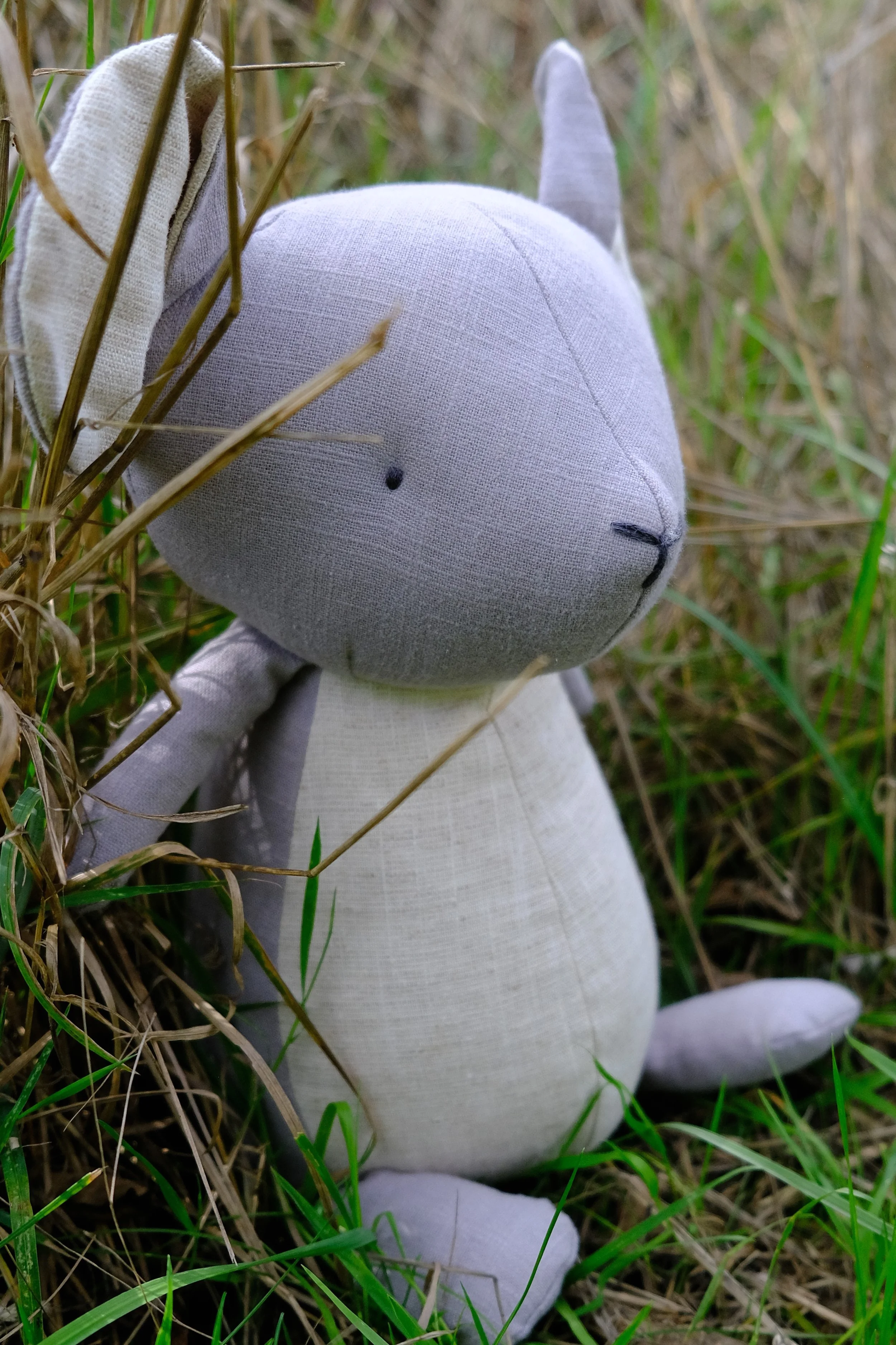 A fabric stuffed toy resembling a cat sitting among tall grass and plants, with a simple face and pointed ears.