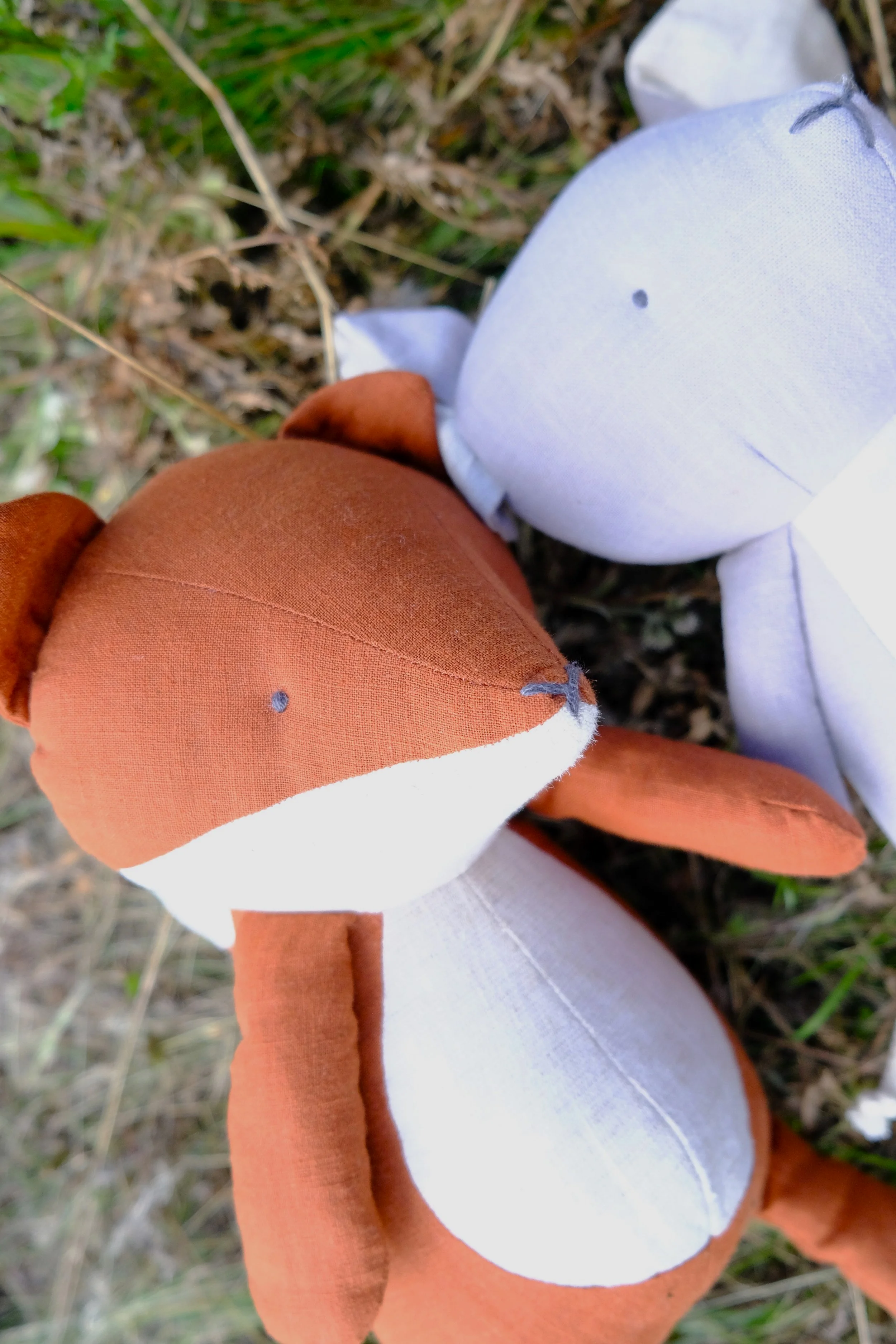 Two stuffed animal toys, one brown and white fox and one white bear, lying on grass and leaves outdoors.