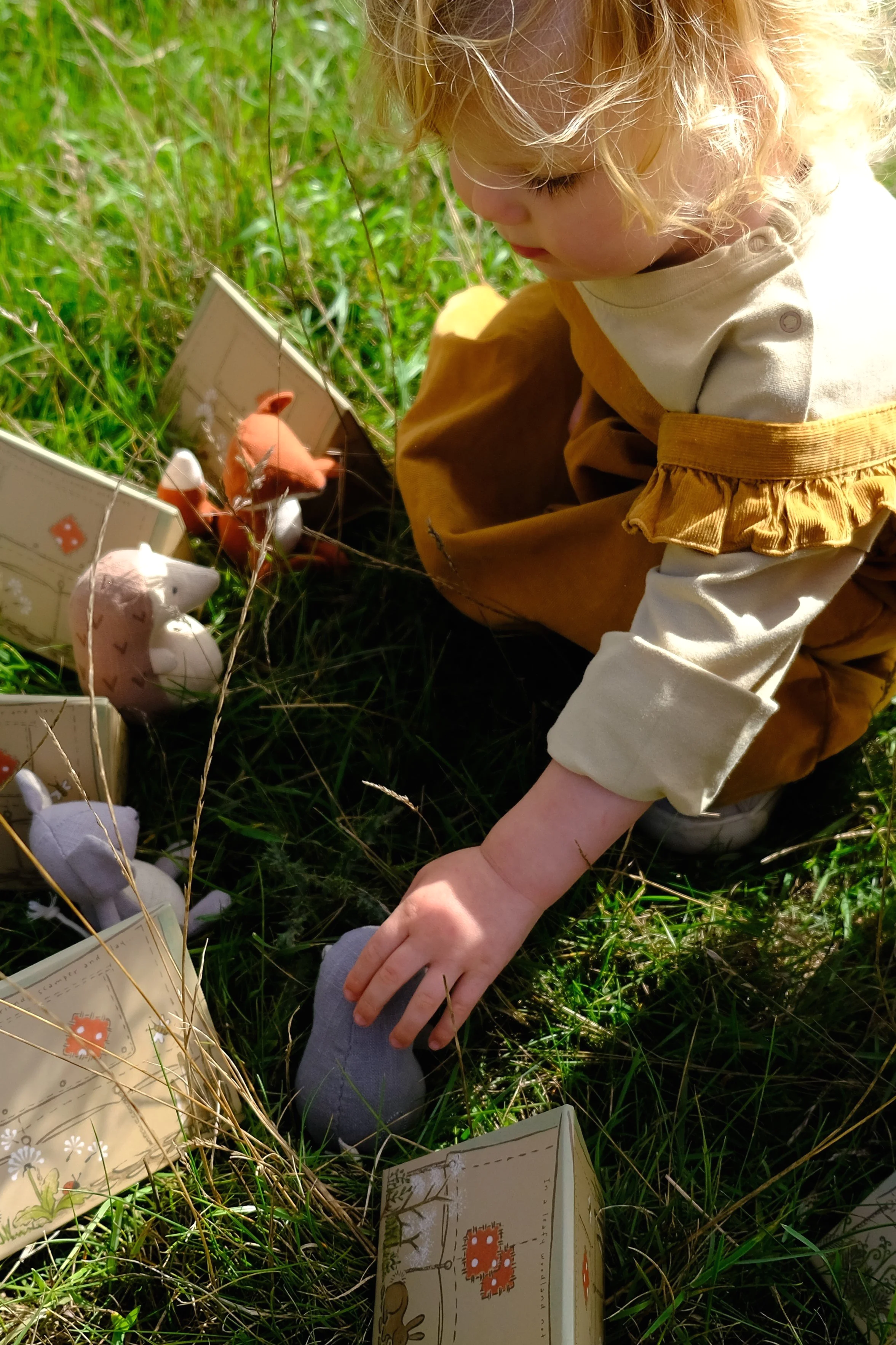 A young child with blonde curly hair playing outdoors in the grass, surrounded by small toy animals and decorated boxes.