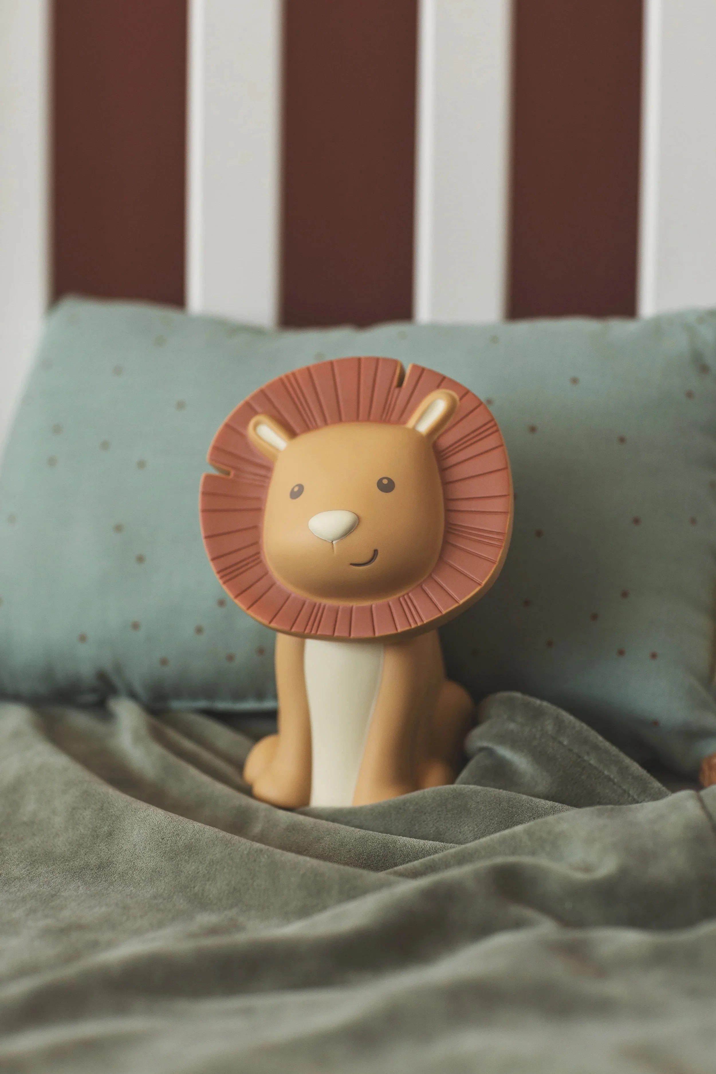 A small lion figurine with a smiling face and a mane, sitting on a bed with gray sheets and a light blue pillow behind it.