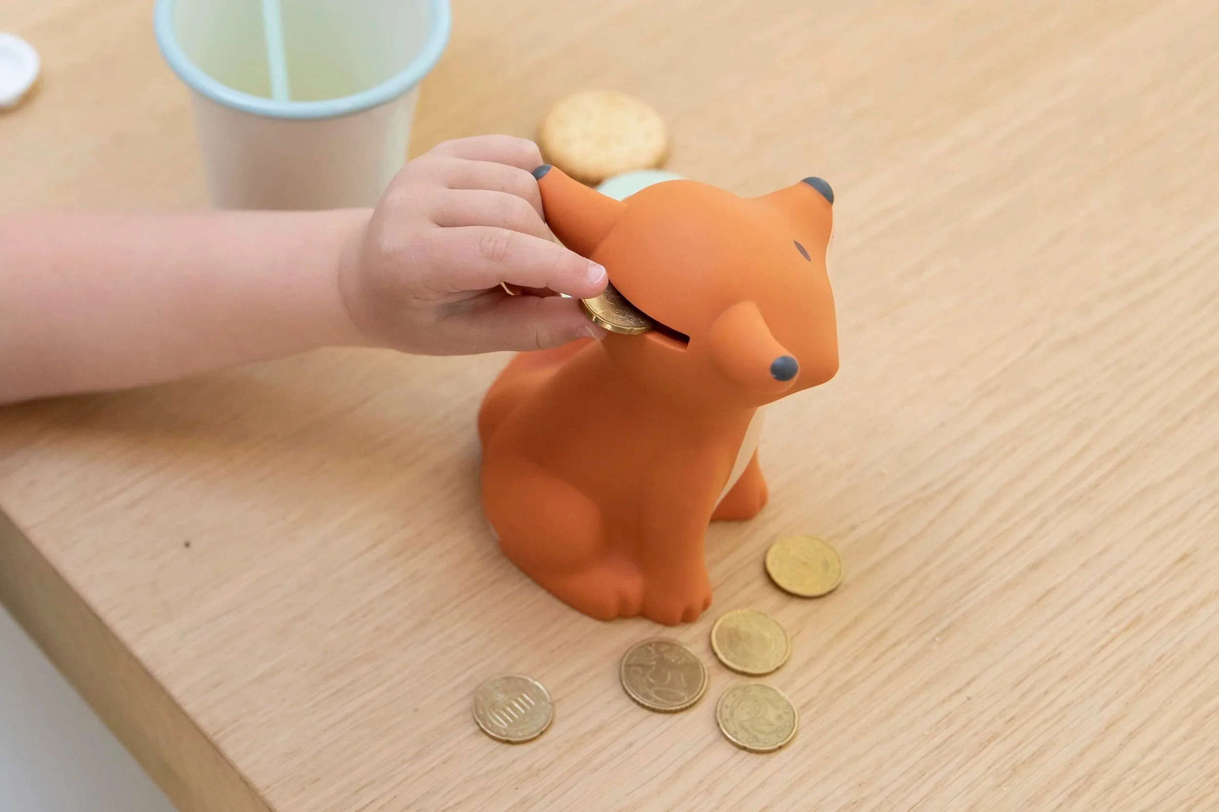 Child's hand inserting a coin into an orange fox-shaped piggy bank on a wooden table with coins around it.