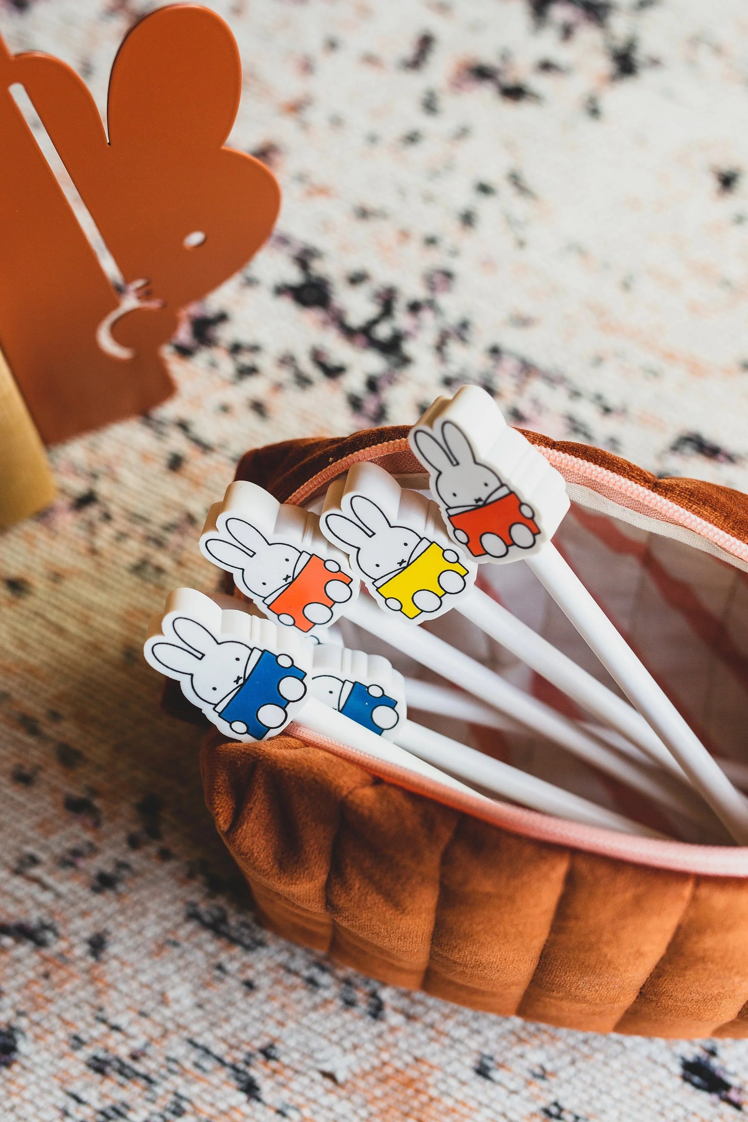 Set of five bunny-themed pens with different colored boxes (red, yellow, blue, orange, and black), placed in a small brown pouch on a patterned rug.